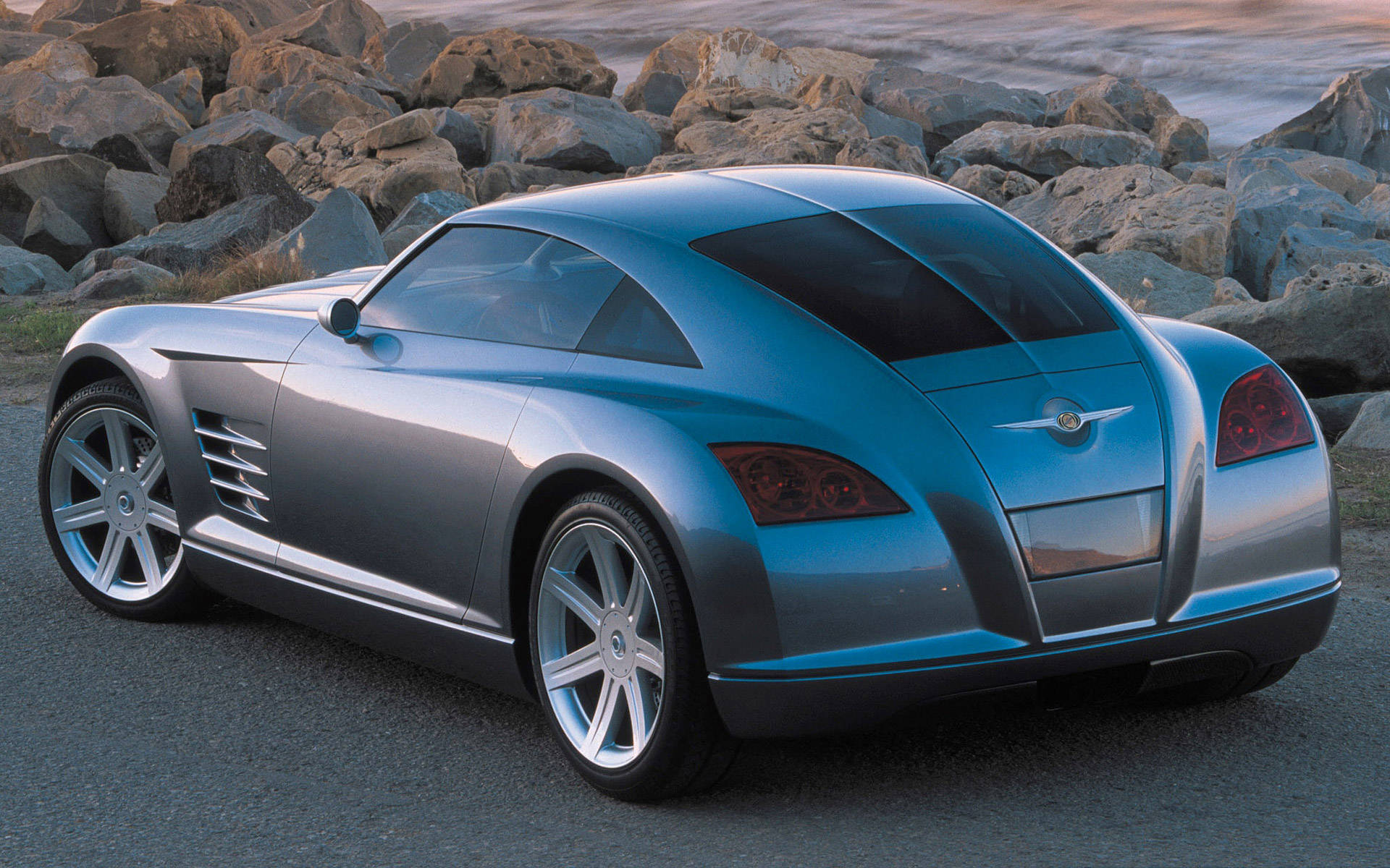 2004 Chrysler Crossfire 動感優雅 壁紙 — 一輛時尚的銀色跑車，帶有紅色尾燈，在日落時停泊在水邊大岩石旁的瀝青路面上。