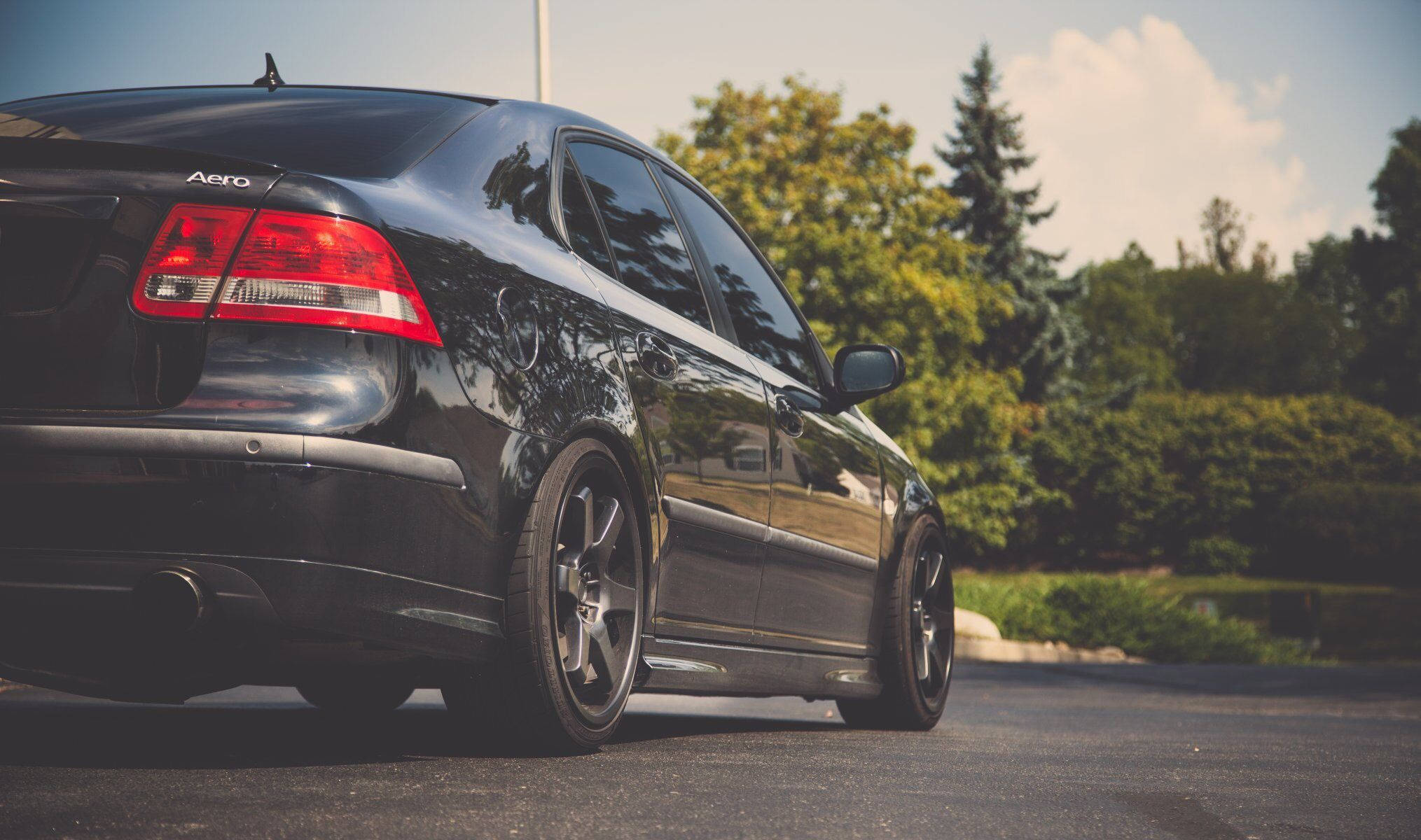 2006 Saab 9-3 Aero 壁紙 — 一輛時尚的黑色轎車配備定制輪圈，停在瀝青路面上，背景是明亮的天空和綠樹。