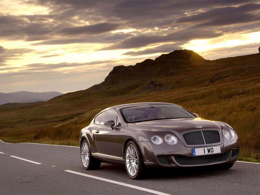 2007年賓利Continental GT運動車 壁紙 — 一輛時尚的灰色賓利 Continental GT 在戲劇性的金色日落天空下沿著蜿蜒的山路巡航。