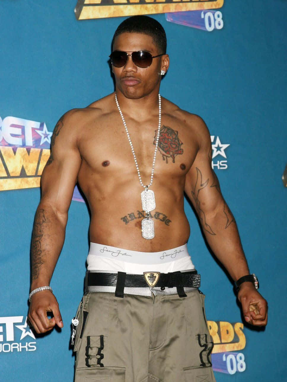 2008 BET 頒獎禮上無上衣的 Nelly 壁紙 — 一位赤膊、紋身、戴著飛行員太陽眼鏡的肌肉男在2008年BET頒獎典禮上自信地擺姿勢，背景為藍色。