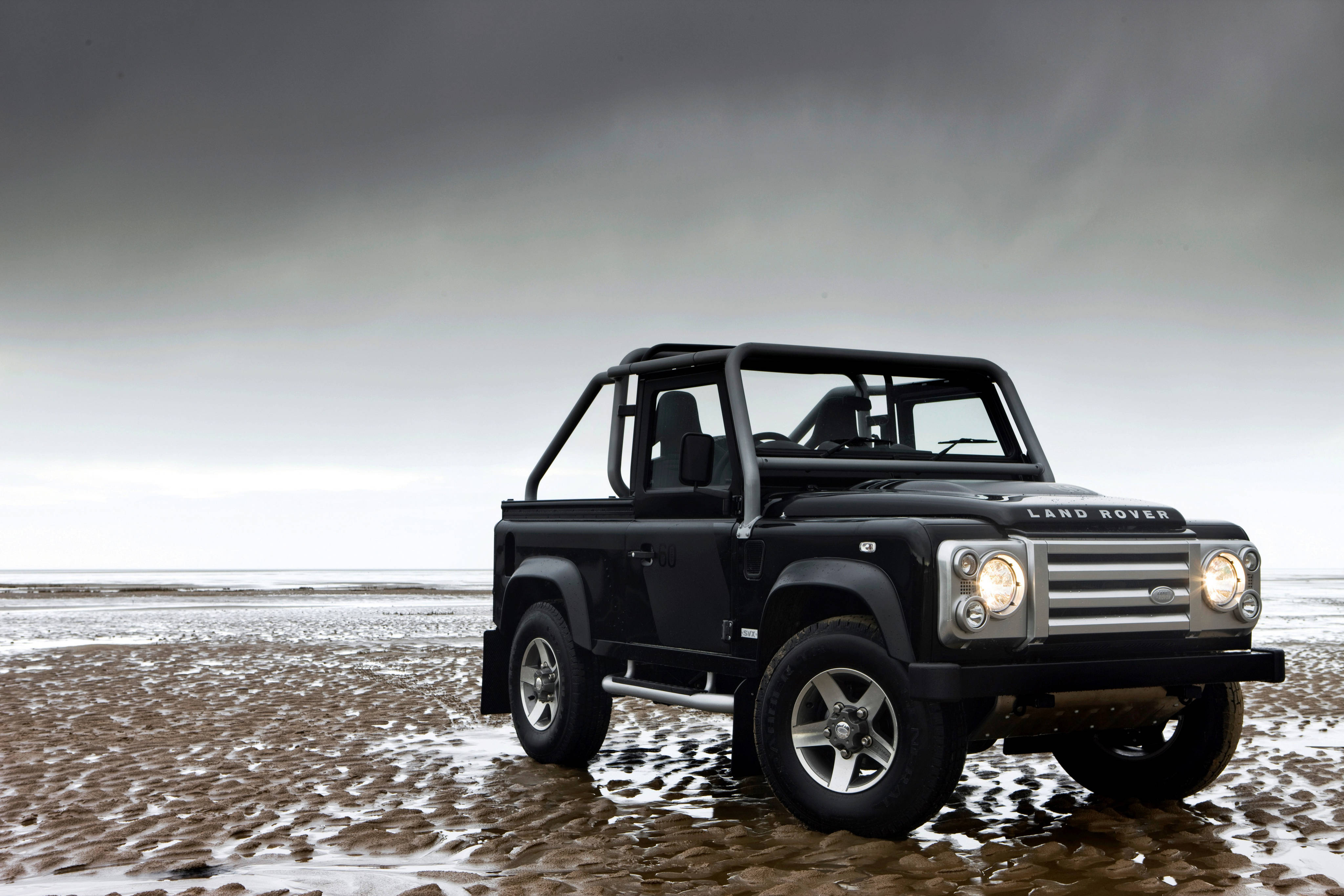2008款黑色路虎 iPhone 主題壁紙 壁紙 — 一輛黑色 Land Rover Defender 在沉重的灰色天空下，堅固地停在濕漉漉的沙灘上。
