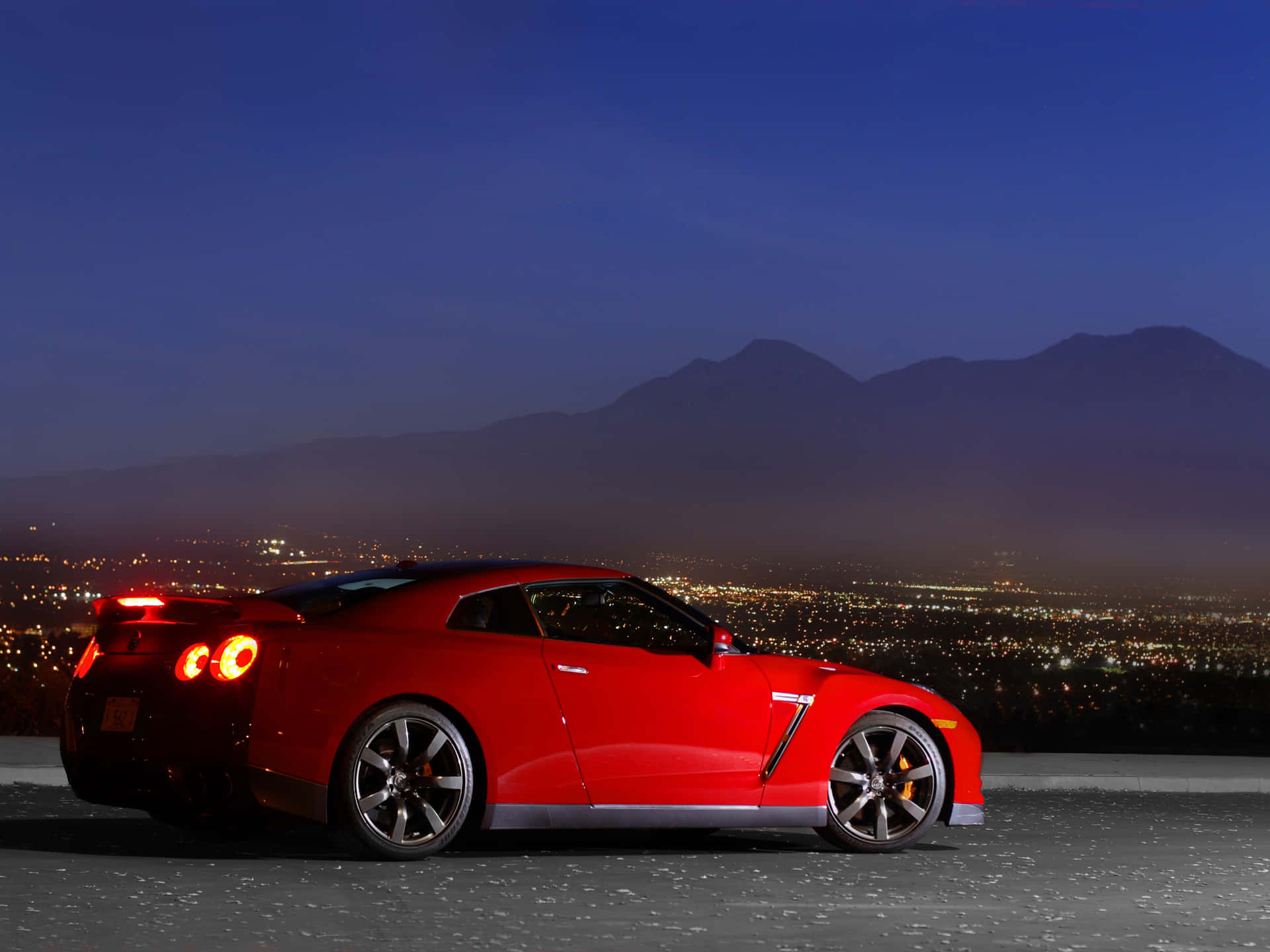 2009 年紅色 Nissan R35 GT-R 車款 壁紙 — 一輛鮮紅色的跑車在深藍色天空下，以遠山為背景，在暮色城市景觀中閃耀。
