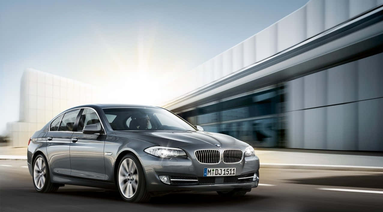 2010 年款 BMW 5 系列 (F10) 深灰 1080p 背景 壁紙 — 一輛時尚的銀色寶馬轎車在明亮的陽光下飛馳而過現代玻璃建築。