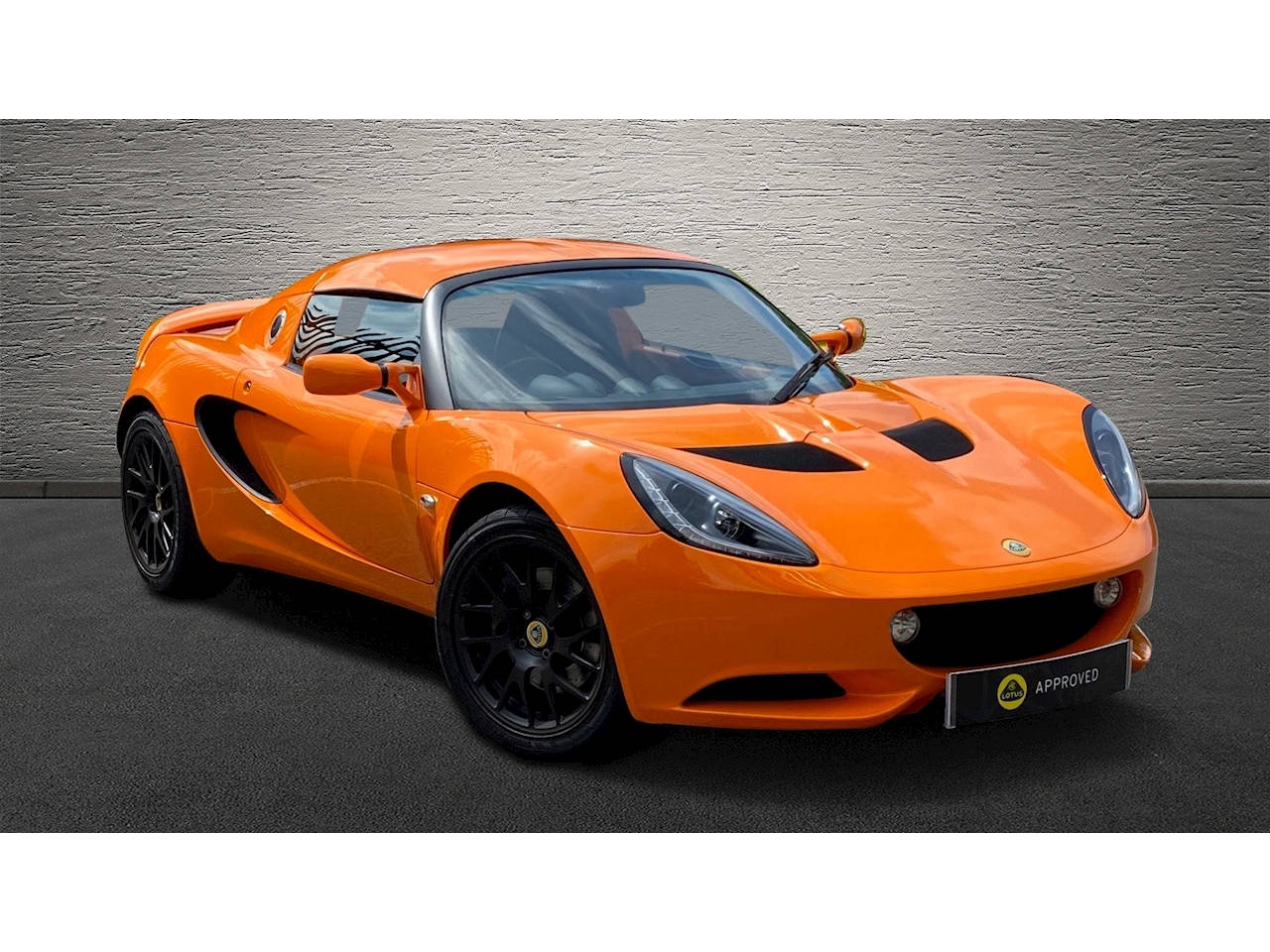 2010款橙色路特斯Elise跑車 壁紙 — 一輛鮮豔的橙色蓮花Elise跑車，帶有黑色飾邊和賽車條紋，停放在有紋理的灰色牆壁前。