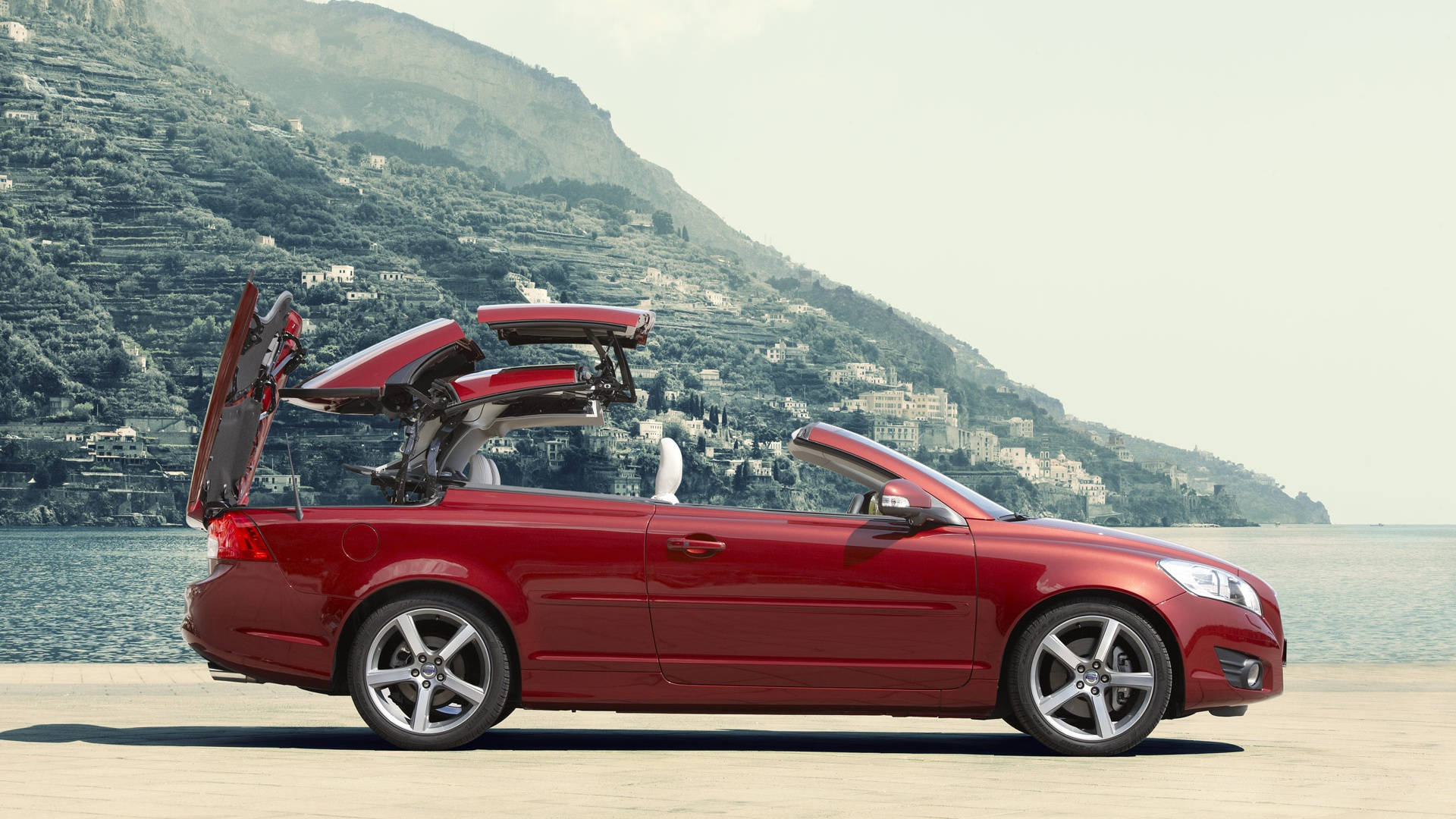2011 沃爾沃 C70 T5 iPhone 壁紙 — 一架鮮紅色開篷跑車，車頂打開，車門升起，停泊在湖邊碼頭，背景是梯田狀的山丘，點綴著房屋。