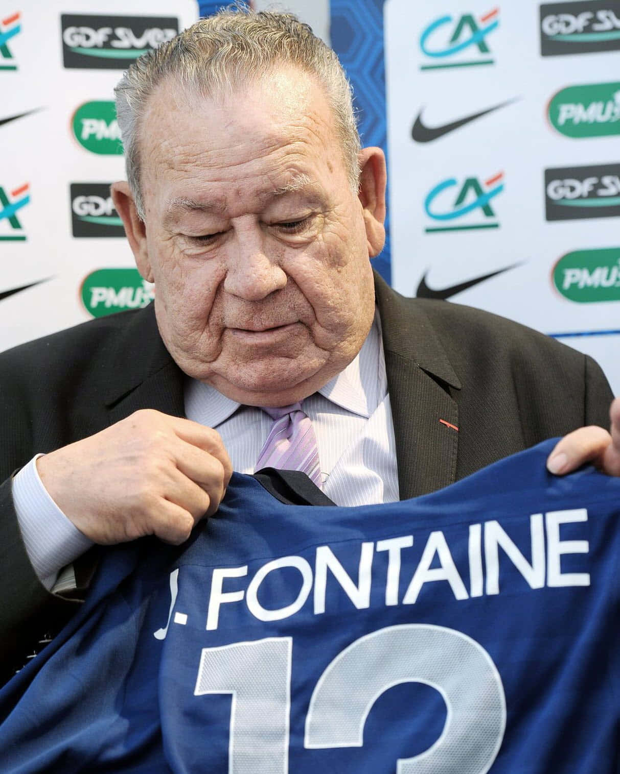 2011年法國足球員Just Fontaine 壁紙 — 一位身穿西裝的長者在記者會上舉起一件印有「J FONTAINE 12」的藍色足球球衣。