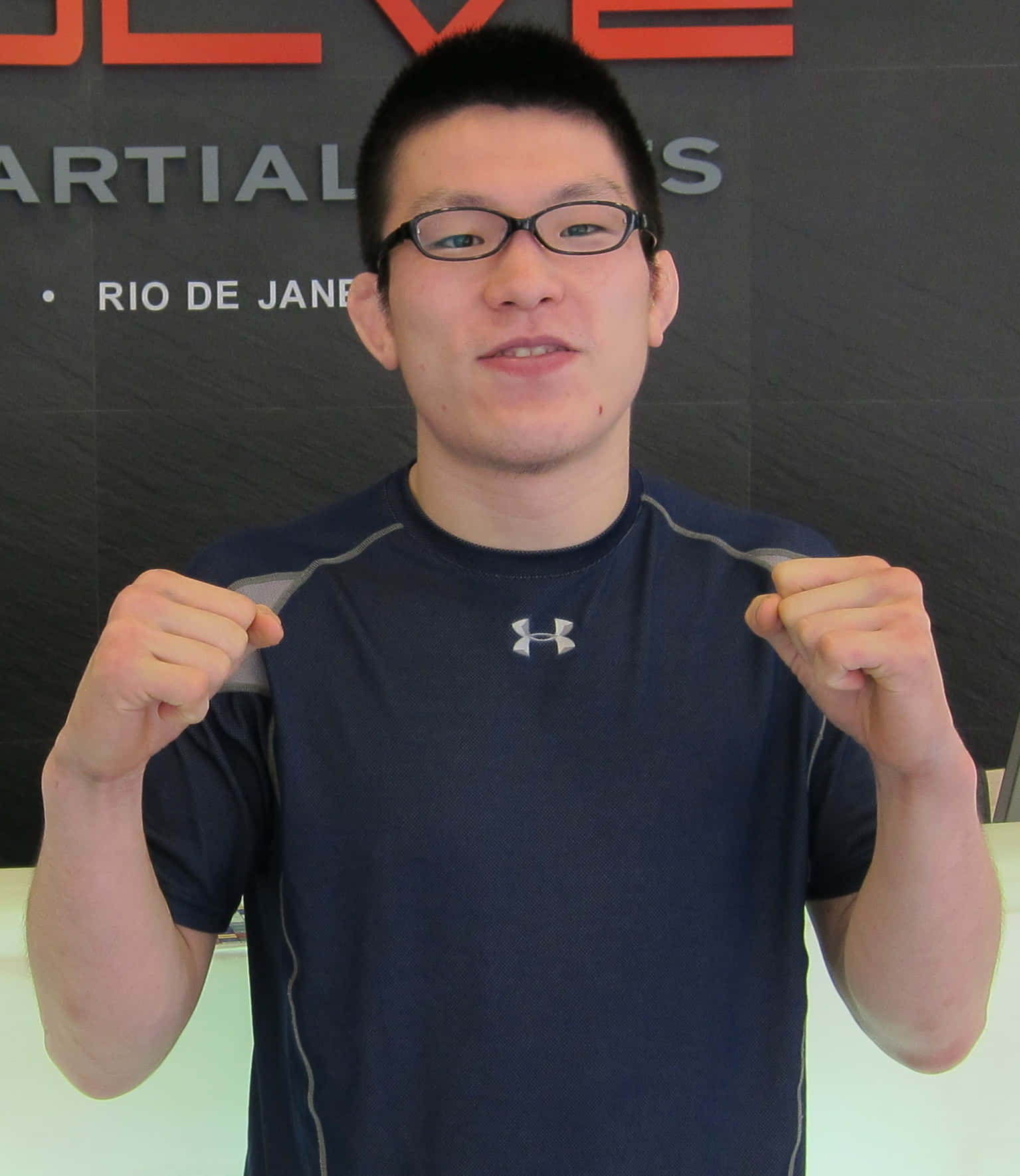 2011年青木真也身穿Evolve MMA戰衣展現霸氣 壁紙 — 一位戴眼鏡、身穿海軍藍Under Armour襯衫的年輕男子在武術工作室的牆前舉起拳頭擺姿勢。