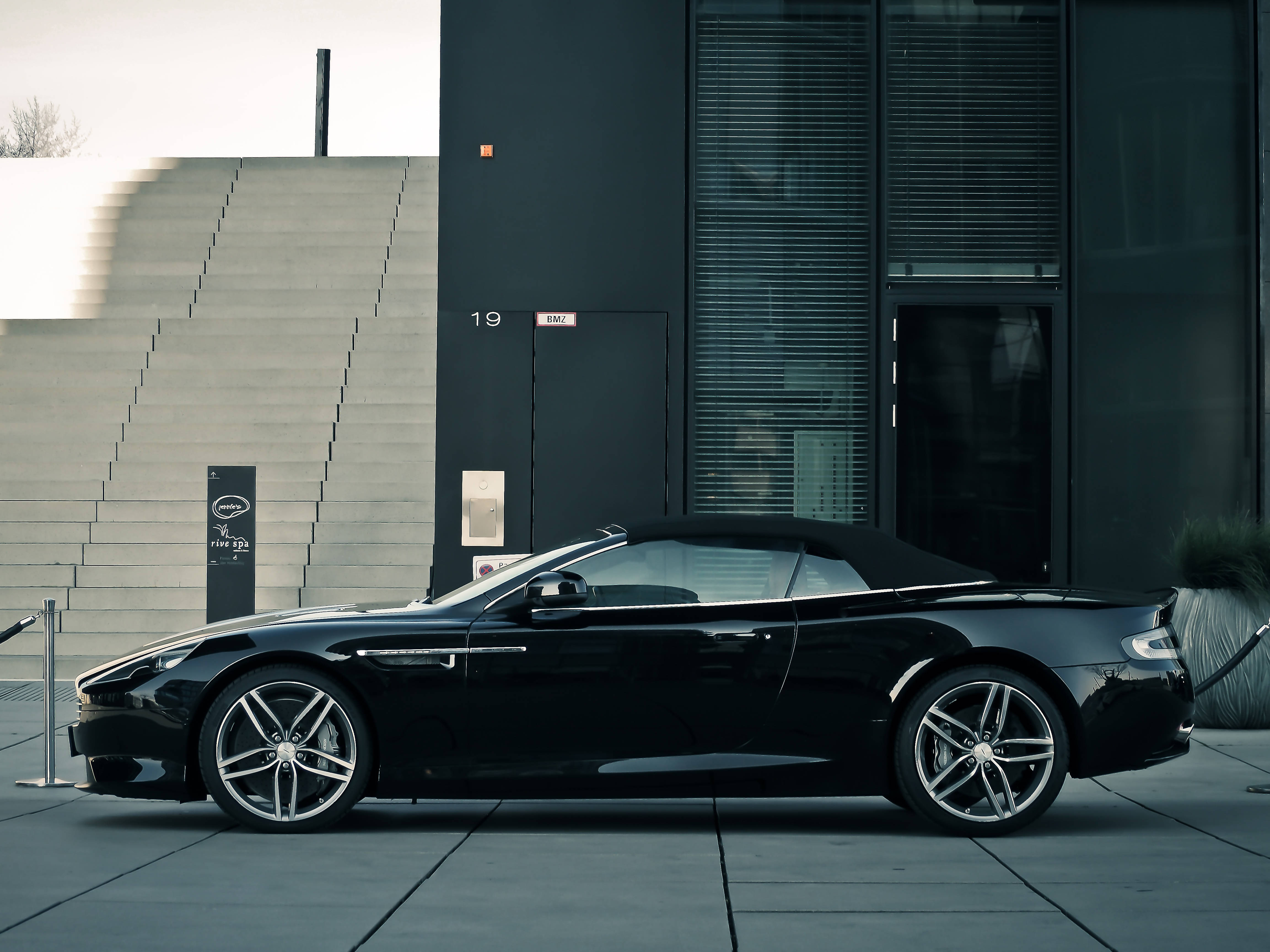 阿斯頓·馬丁 2012 碳纖維 DBS Volante 壁紙 — 一架時尚的黑色Aston Martin開篷車停泊在現代建築前，建築物有玻璃窗和混凝土階梯。