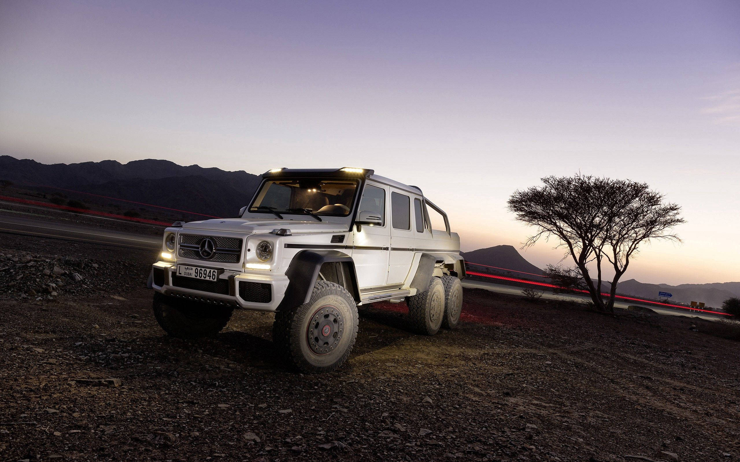 2013 Mercedes-Benz G63 AMG 壁紙 — 一輛白色平治 G63 AMG 6x6 在黃昏時停在碎石上，車流燈光劃過，孤樹襯托紫色山巒。