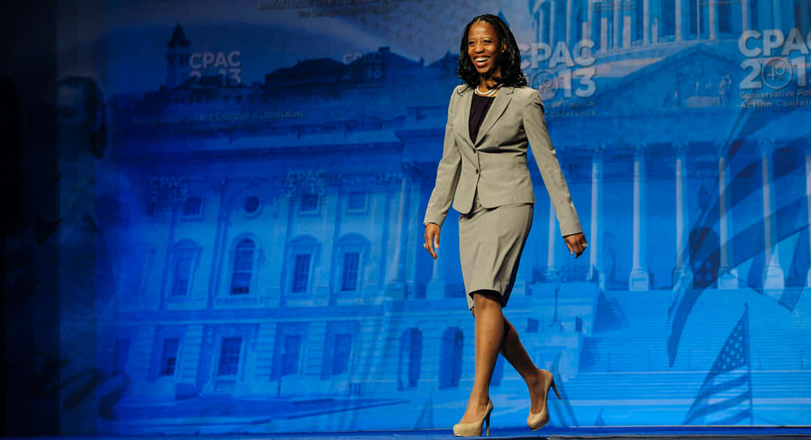 米婭·洛夫出席2013年CPAC 壁紙 — 一位微笑的灰色西裝女士自信地走在舞台上，背景是帶有政府建築的藍色調CPAC 2013背景。