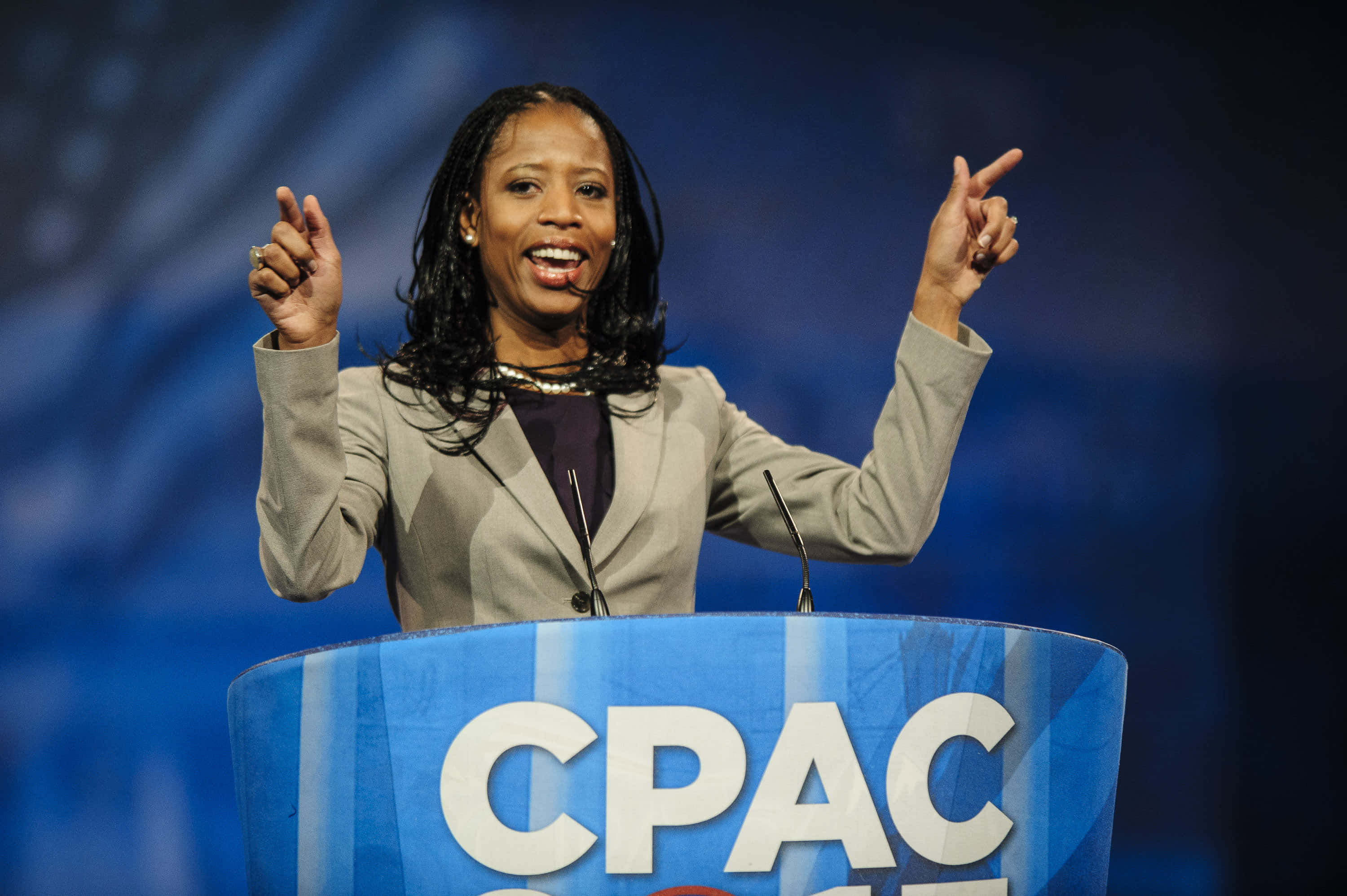 米亞·洛夫於2013年CPAC講台演講 壁紙 — 一位身穿米色西裝外套的女性在藍色背景燈光的CPAC講台上熱情演講。