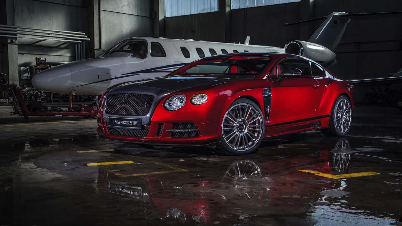 2013款紅色賓利Continental GT背景圖 壁紙 — 一輛亮紅色的Mansory Bentley Continental GT在潮濕的機庫中，於工業燈光下與私人飛機並排閃耀。