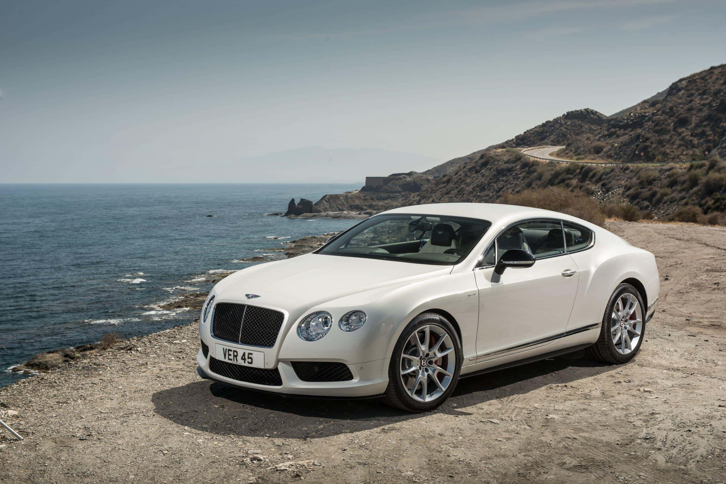 2014 賓利 Continental GT Sport 壁紙 — 一架純白色的賓利 Continental GT 停泊在陽光明媚的懸崖上，俯瞰深藍色的海洋，背景是崎嶇的山丘。