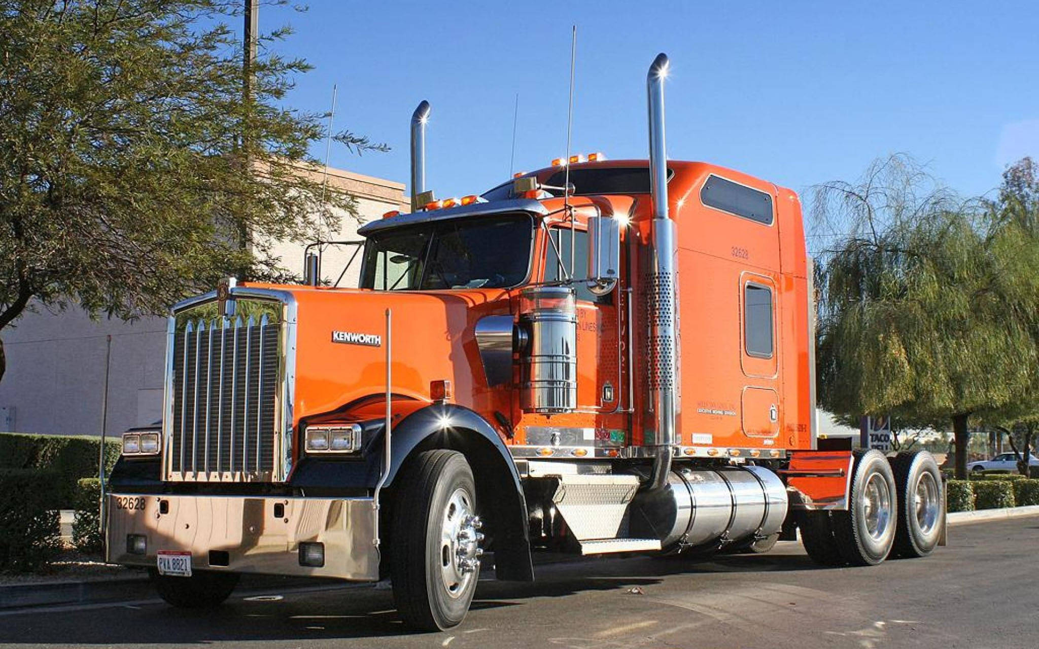 2014年肯沃斯W900 壁紙 — 一輛亮橙色的Kenworth半掛卡車在晴朗的藍天下，停泊在陽光普照、綠樹成蔭的街道上閃閃發光。
