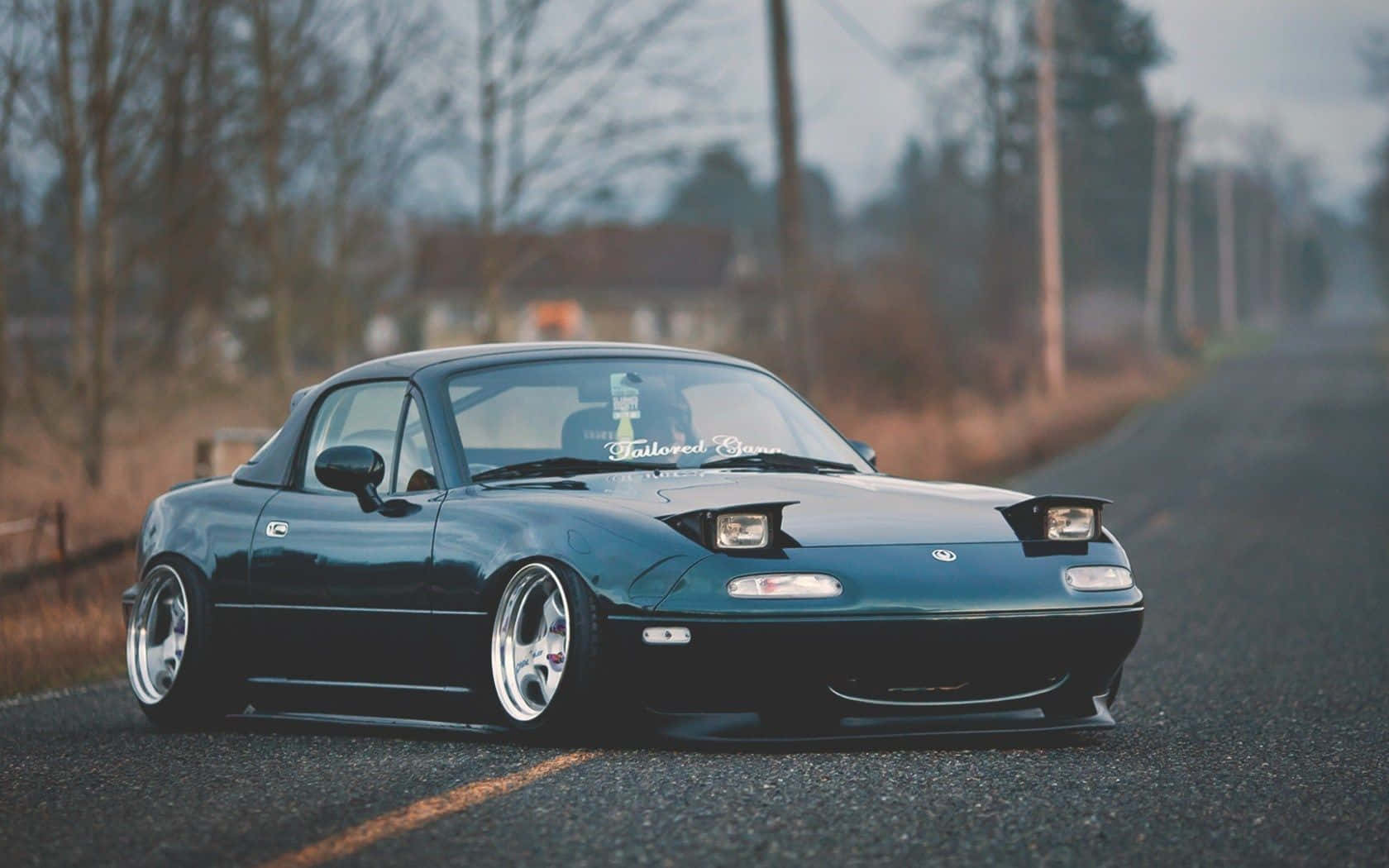 2014年馬自達MX-5 Miata跑車美學攝影 壁紙 — 一架時尚深青綠色的改裝Miata跑車，配備定制輪圈，在黃昏時低趴於空曠的鄉村道路上。