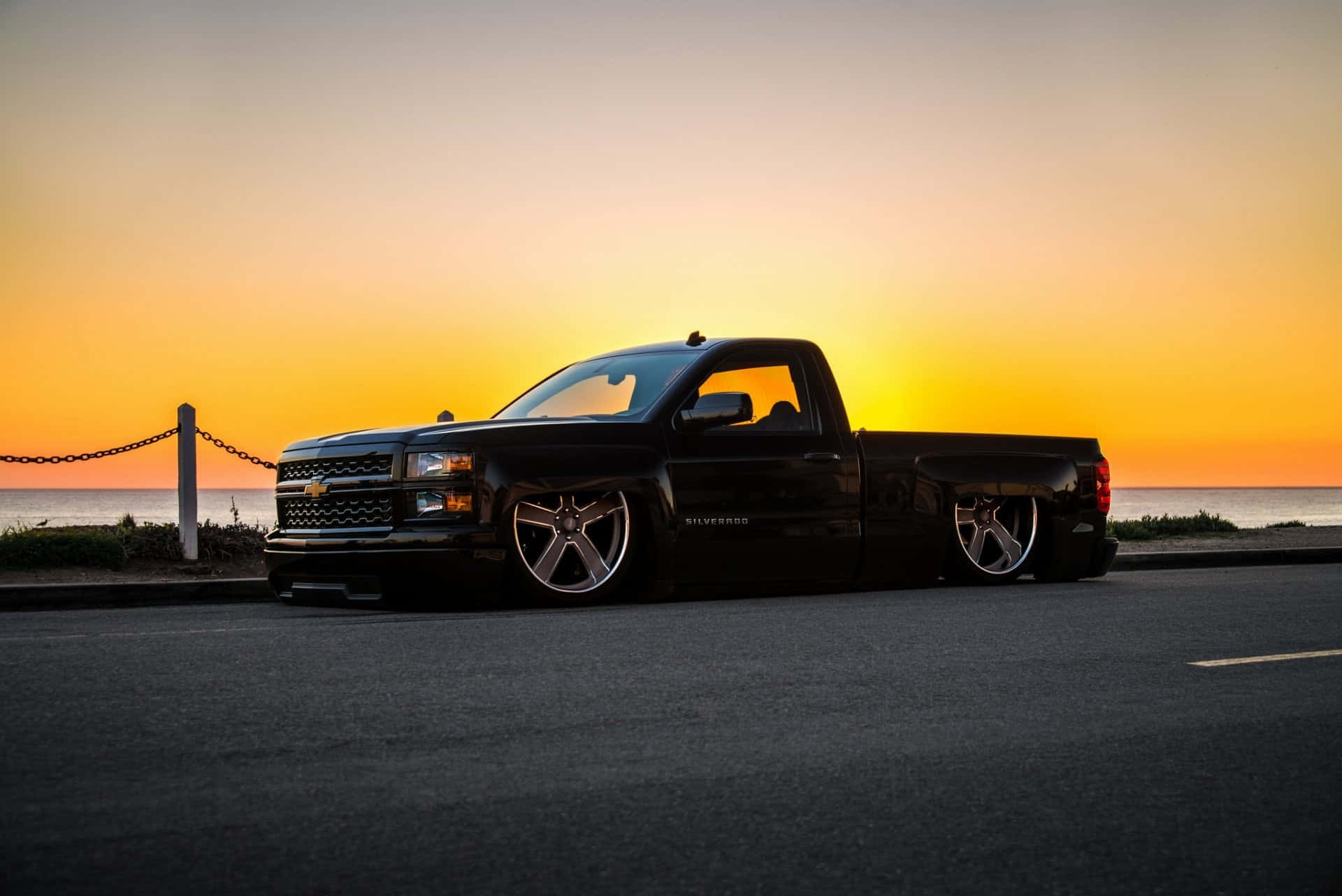 2014款雪佛蘭Silverado 1500皮卡日落攝影 壁紙 — 一架時尚黑色降低車身的雪佛蘭 Silverado 皮卡車停泊在日落時分的海岸公路上，天空呈現鮮豔的橙色和黃色。
