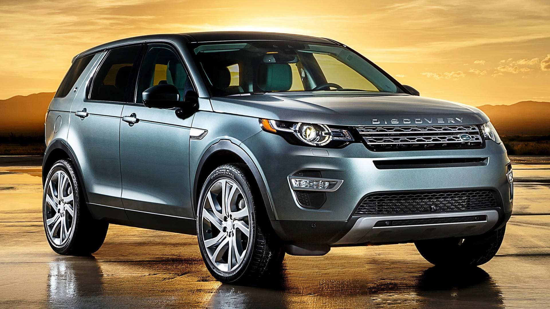 2015 路虎發現運動版 iPhone 主題壁紙 壁紙 — 一輛時尚的銀色Land Rover Discovery SUV在金色夕陽下閃閃發光，在潮濕的沙漠道路上投下長長的影子。