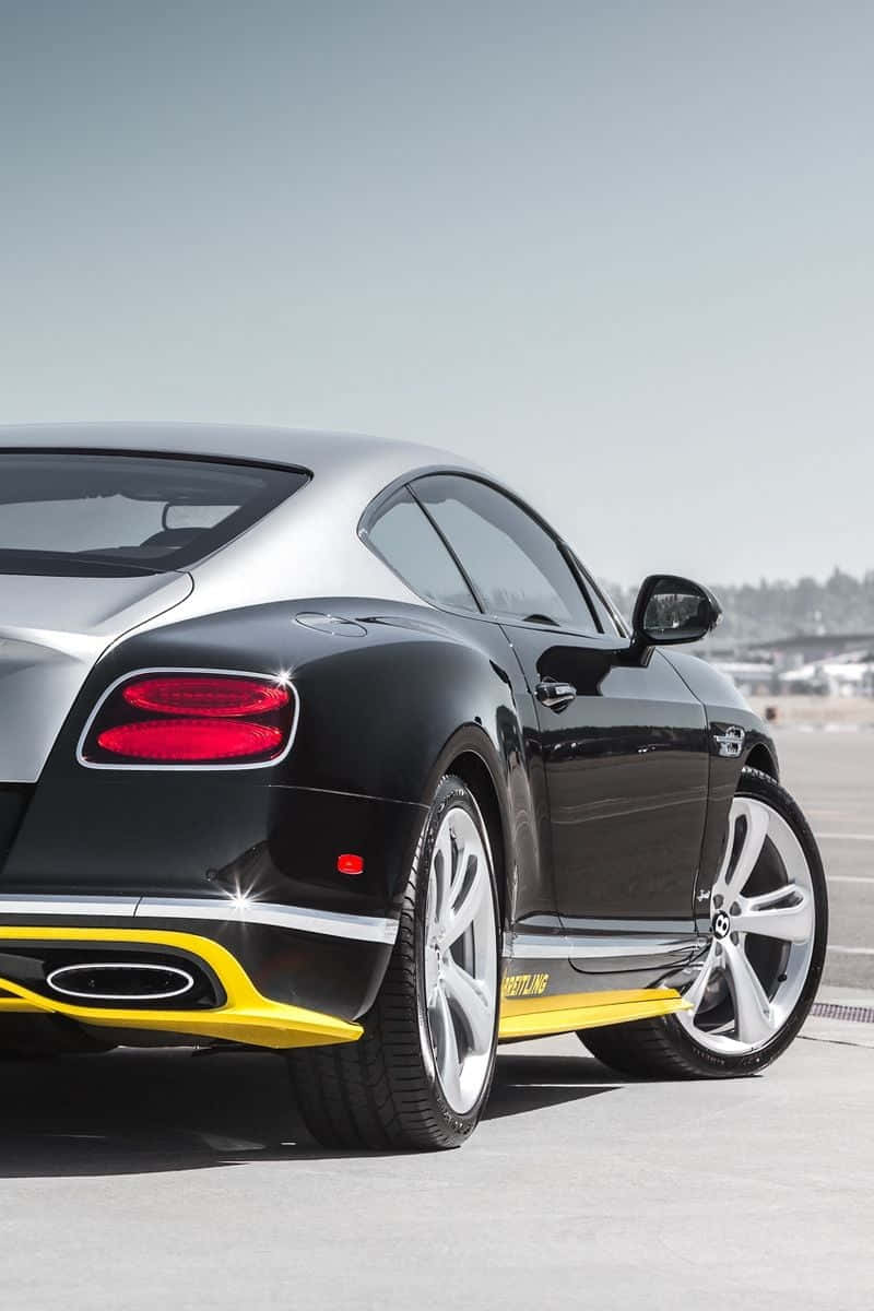 2015款賓利iPhone壁紙 壁紙 — 一輛時尚的黑色賓利 Continental GT 帶有醒目的黃色點綴，在明亮的日光下在鋪砌的路面上閃閃發光。