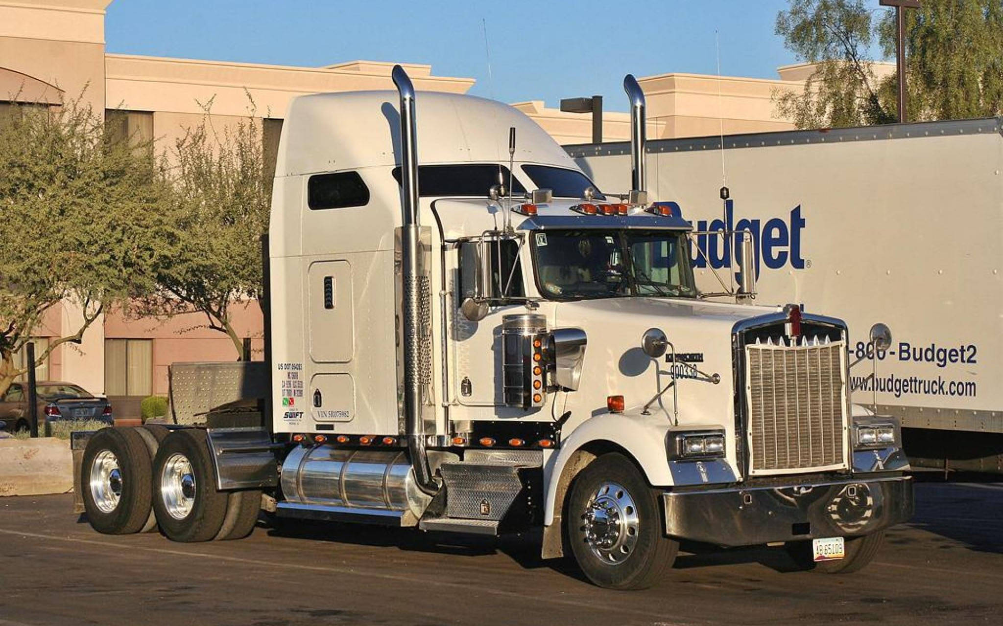 2015年Kenworth W900卡車 壁紙 — 一輛帶有鍍鉻裝飾的純白色Kenworth半掛卡車停泊在陽光普照的停車場，旁邊有一輛Budget拖車。