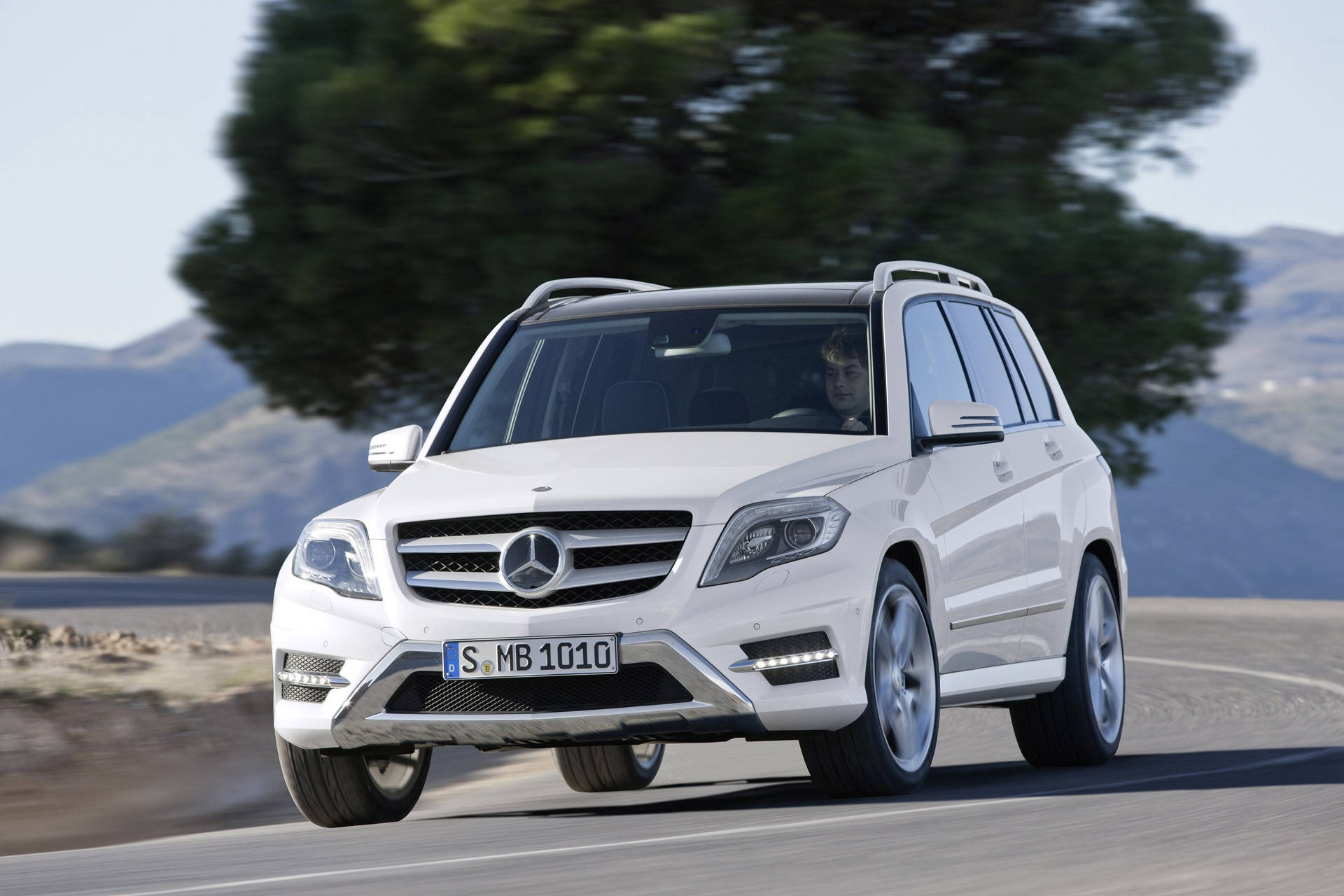 白色Mercedes GLK SUV在山路上行駛，附近有一棵大樹。