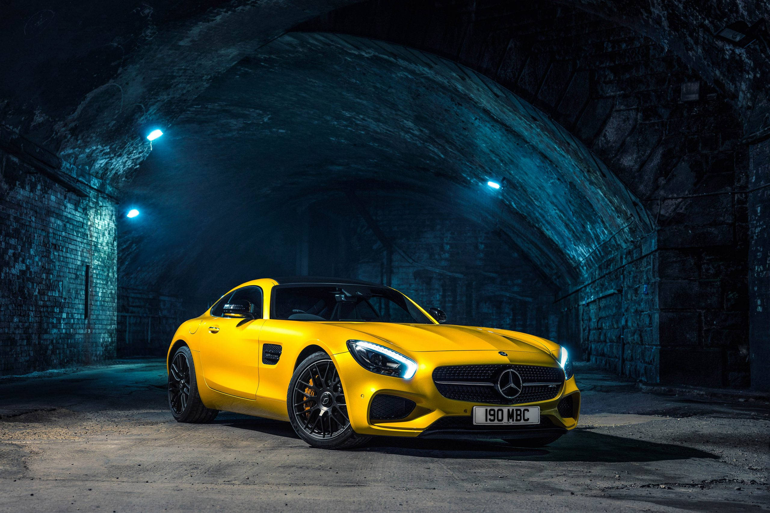 2016款黃色梅賽德斯-奔馳AMG 壁紙 — 一輛鮮黃色的梅賽德斯-AMG GT跑車在黑暗的拱形石隧道中，在藍色聚光燈下閃閃發光。