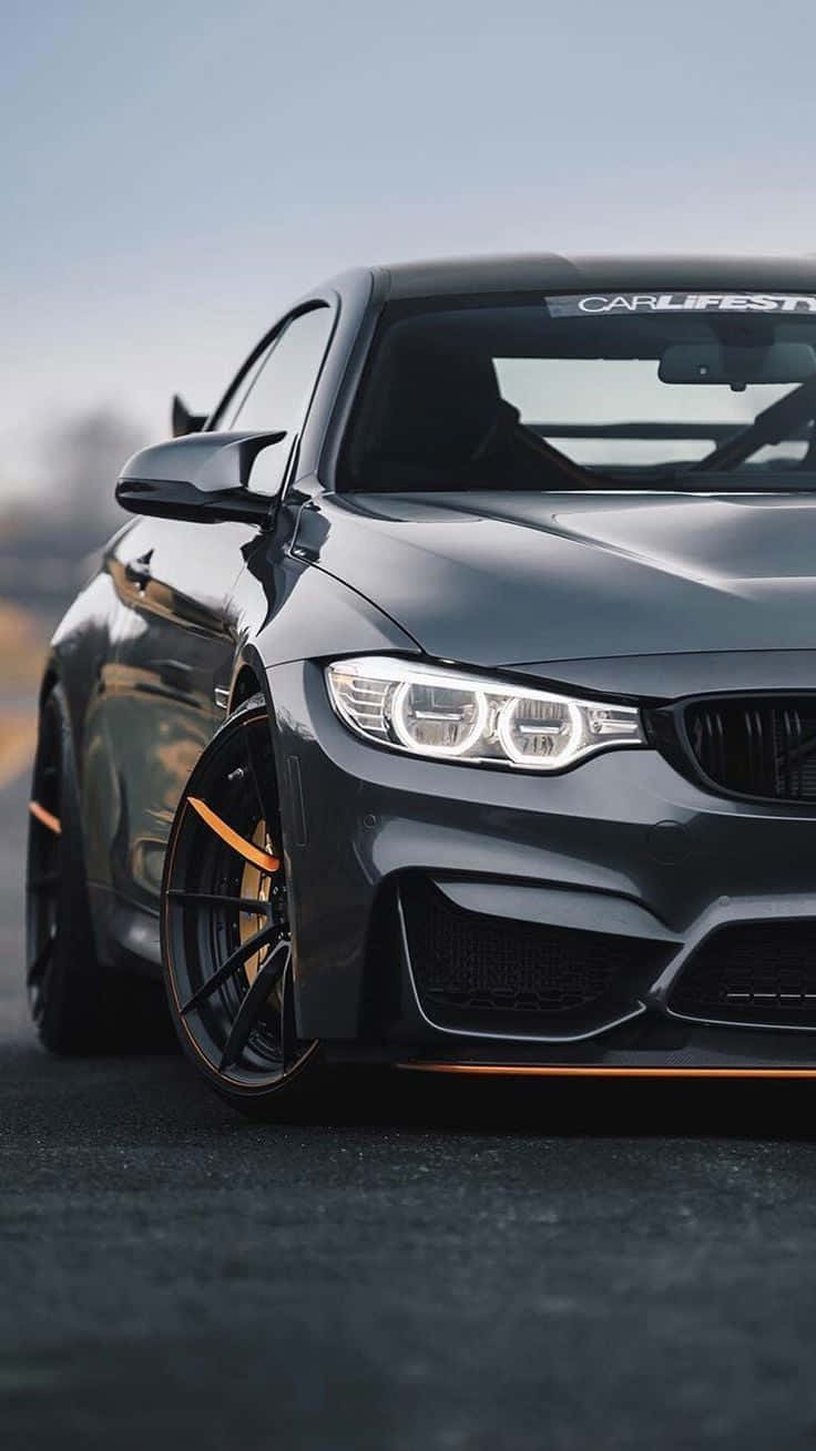 2016 年黑色 Pixel 3 BMW M4 GTS 背景 壁紙 — 一輛時尚的啞光灰色BMW M4，帶有橙色點綴和發光的車頭燈，停在瀝青路面上，處於柔和日光下。