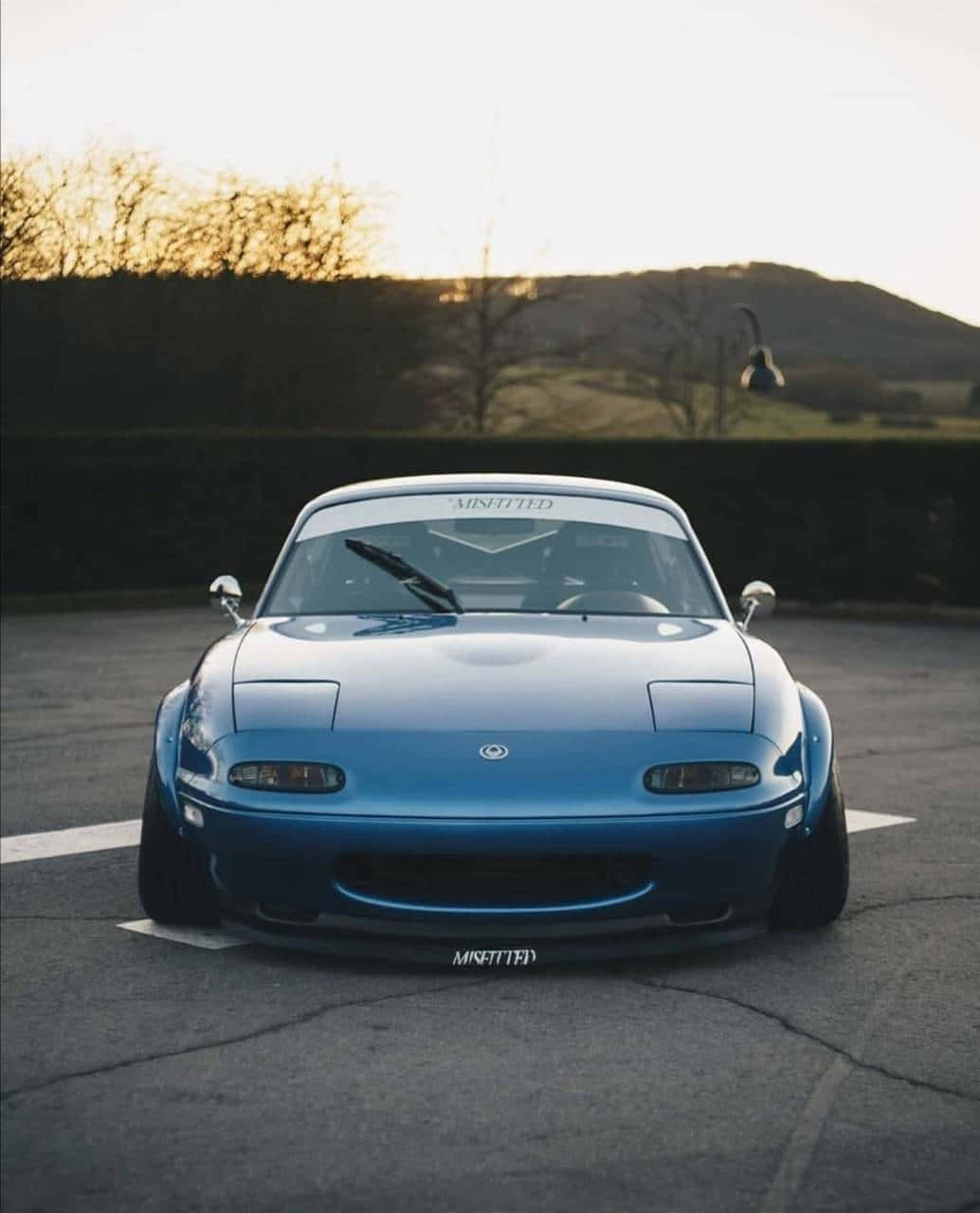 2016年藍色 Mazda MX-5 Miata 前視角度 壁紙 — 一輛充滿活力的藍色萬事得MX-5跑車，降低懸掛，停在夕陽下的瀝青路上，背景有山丘。