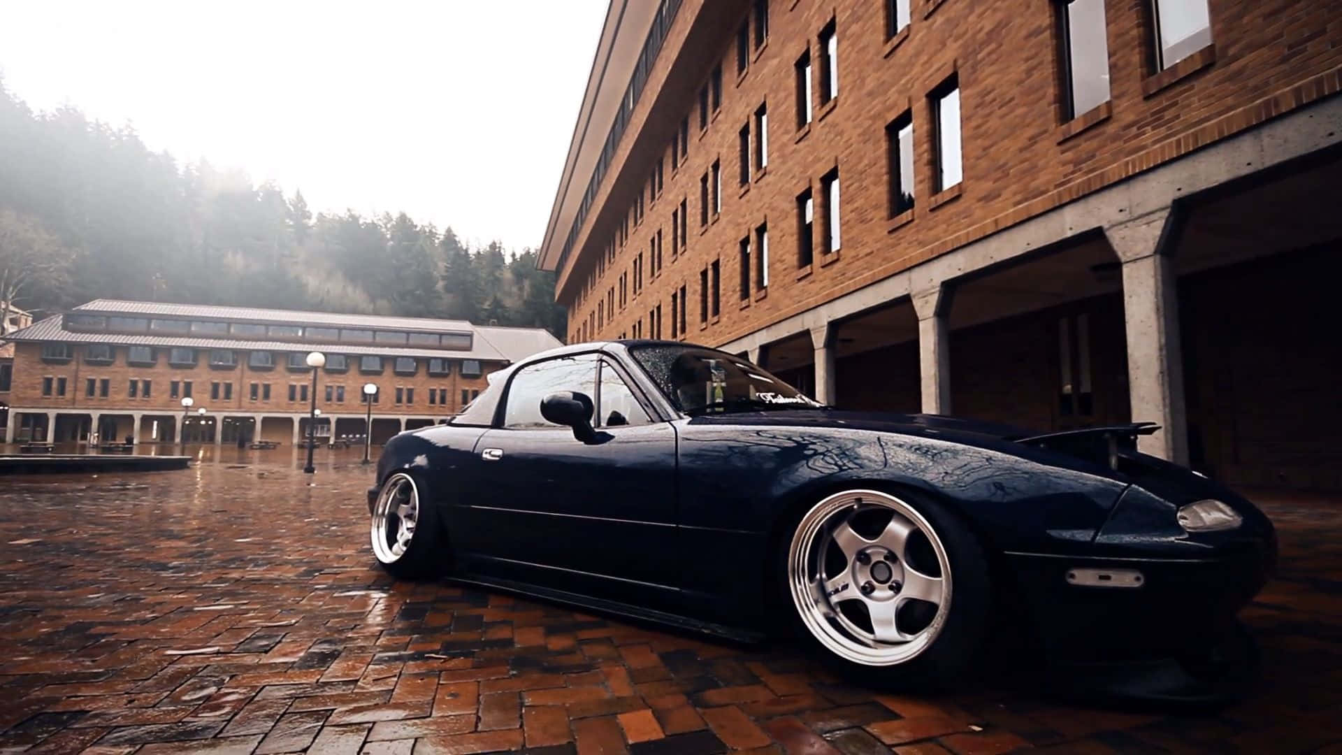 2016年馬自達MX-5 Miata深藍色跑車 壁紙 — 一輛時尚的黑色跑車配銀色輪圈，停在濕磚廣場上，旁邊是磚砌建築，背景有霧氣繚繞的森林山丘。
