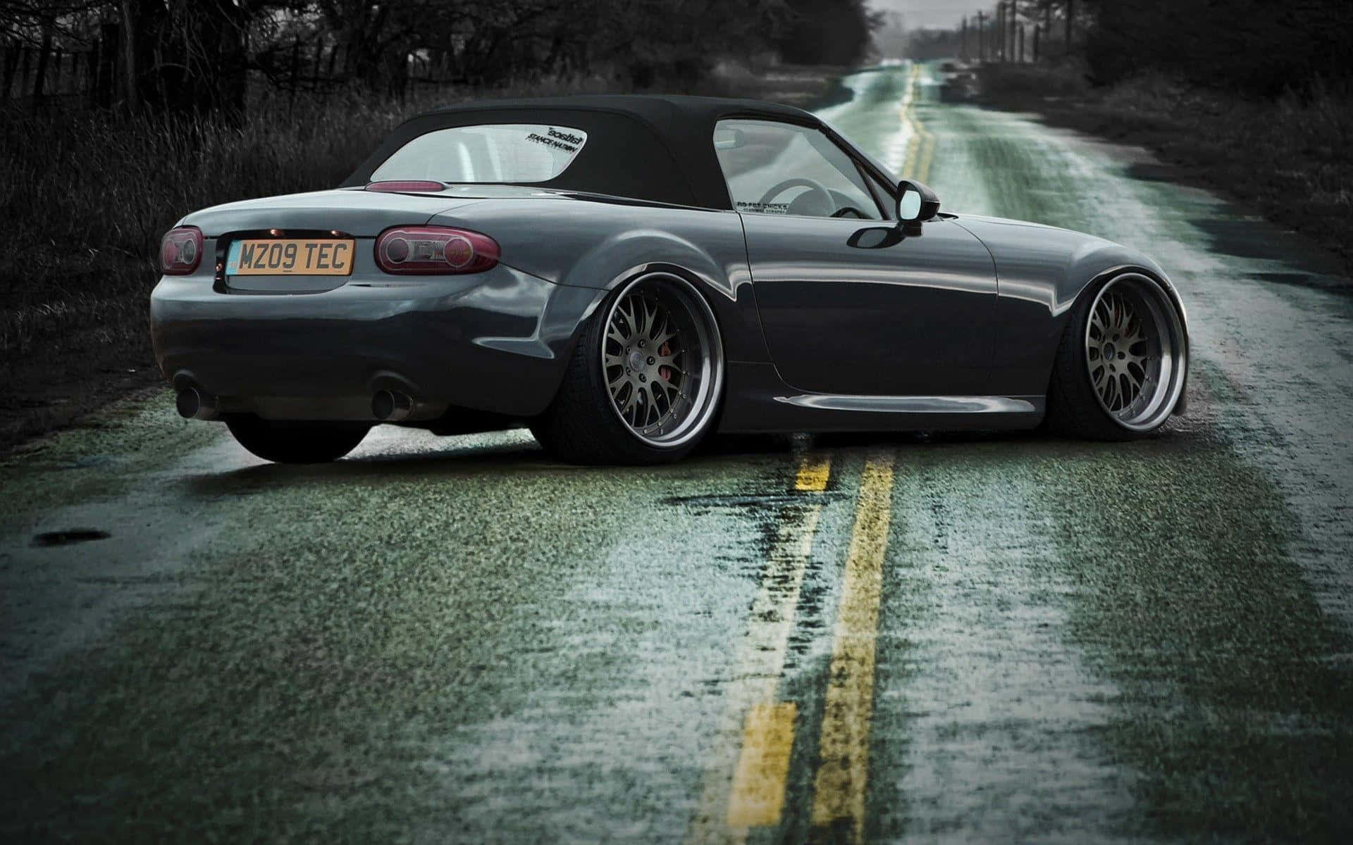 2016年黑色 Mazda MX-5 Miata 跑車 壁紙 — 一架時尚的灰色開篷跑車配備定制輪圈，停泊在潮濕的鄉村道路上，天空多雲。