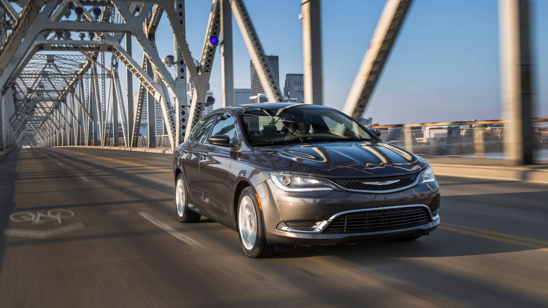 2017 Chrysler 200 黑色車款 壁紙 — 一架時尚的灰色房車在晴朗的藍天下駛過鋼桁架橋，背景是城市天際線。