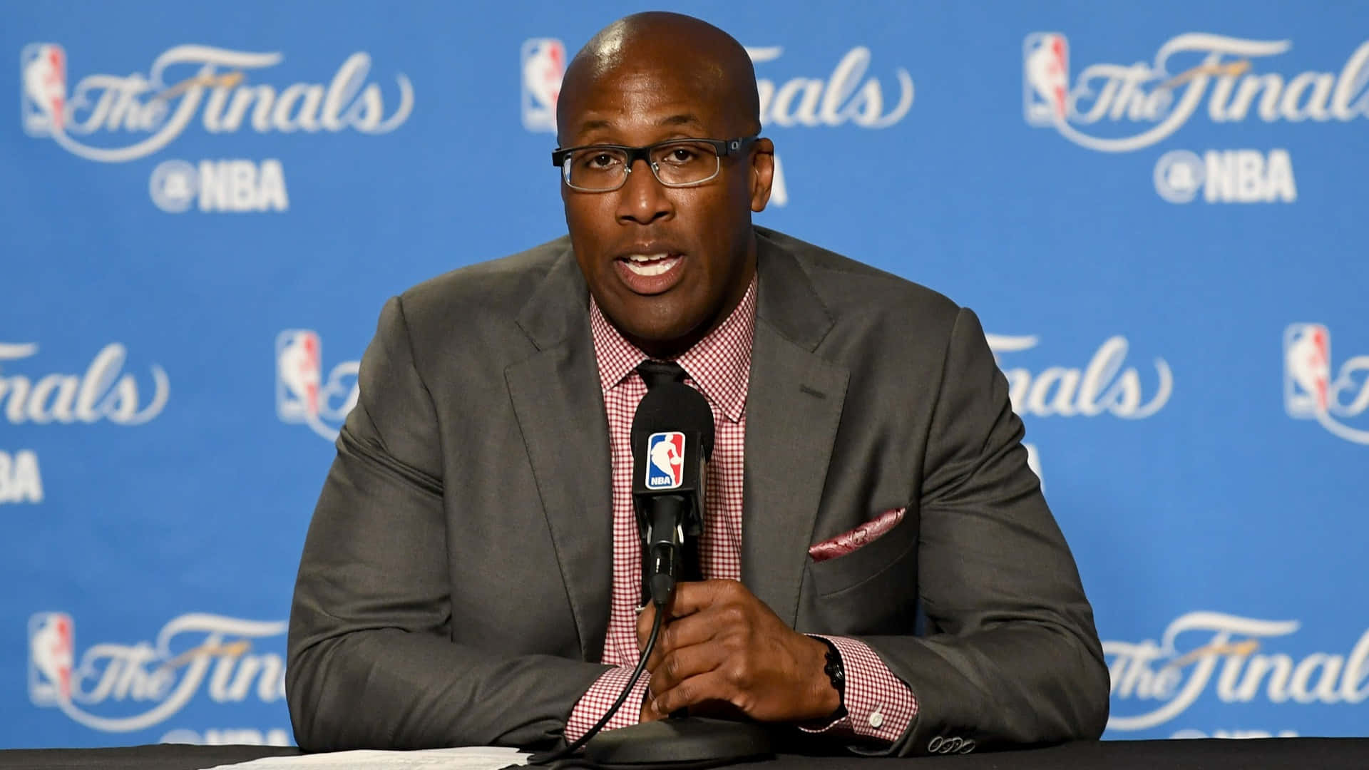 2017年NBA總決賽麥克·布朗教練出席記者會 壁紙 — 一位戴眼鏡、身穿灰色西裝的光頭黑人男子在NBA總決賽記者會上對著麥克風發言。
