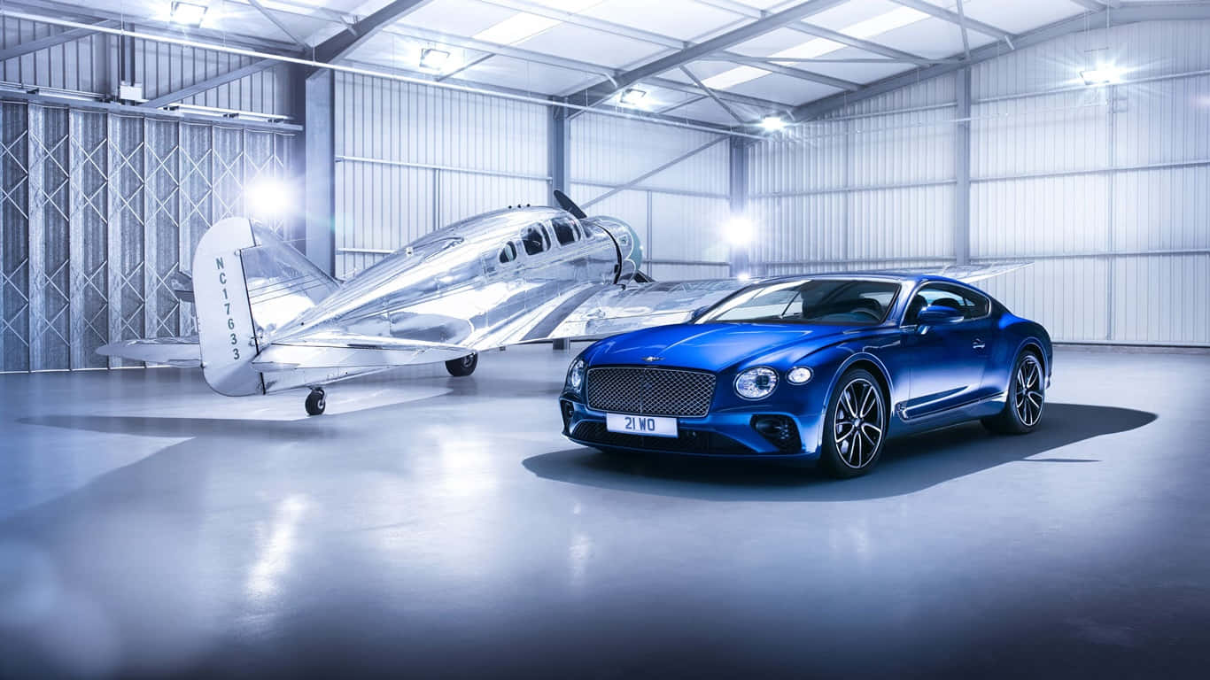 2017款藍色賓利Continental GT背景圖 壁紙 — 一架流線型的藍色賓利 Continental GT 停在一架銀色復古飛機旁邊，位於明亮的工業機庫內。