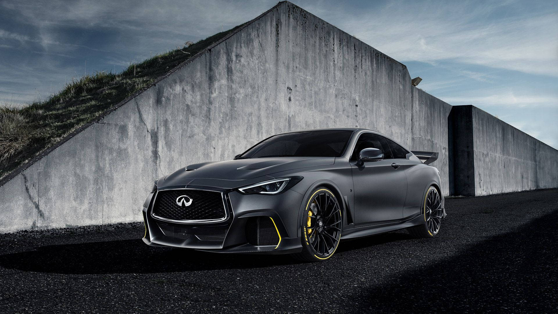2018 英菲尼迪 Q60 壁紙 — 一架啞光灰色英菲尼迪跑車，帶有黃色點綴，停泊在陰天下一座高聳的混凝土牆旁。