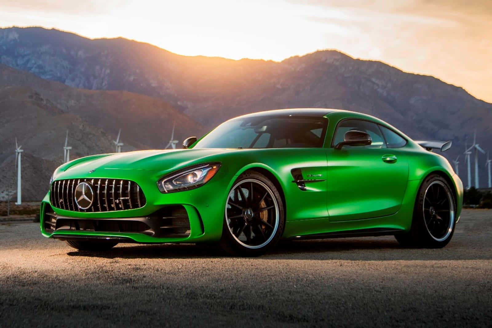 最出色的2018款AMG GT-R（已停產）背景 壁紙 — 一輛充滿活力的綠色 Mercedes-AMG GT 跑車在金色夕陽下閃耀，背景是山脈和風力發電機。