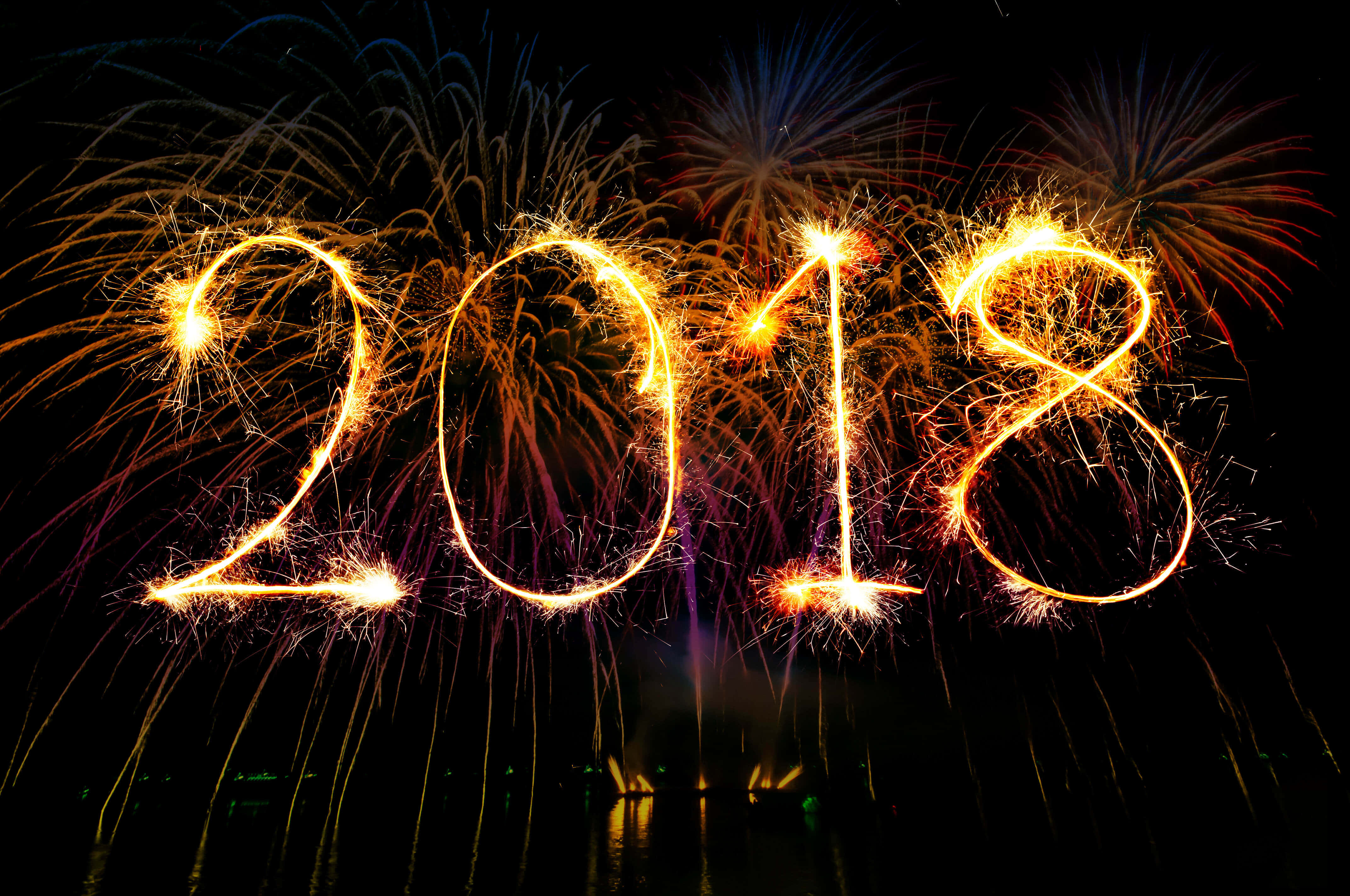 2018 桌布 壁紙 — 2018年以發光的金色煙花棒拼寫，背景是黑暗的夜空，後面有色彩繽紛的煙火爆炸。