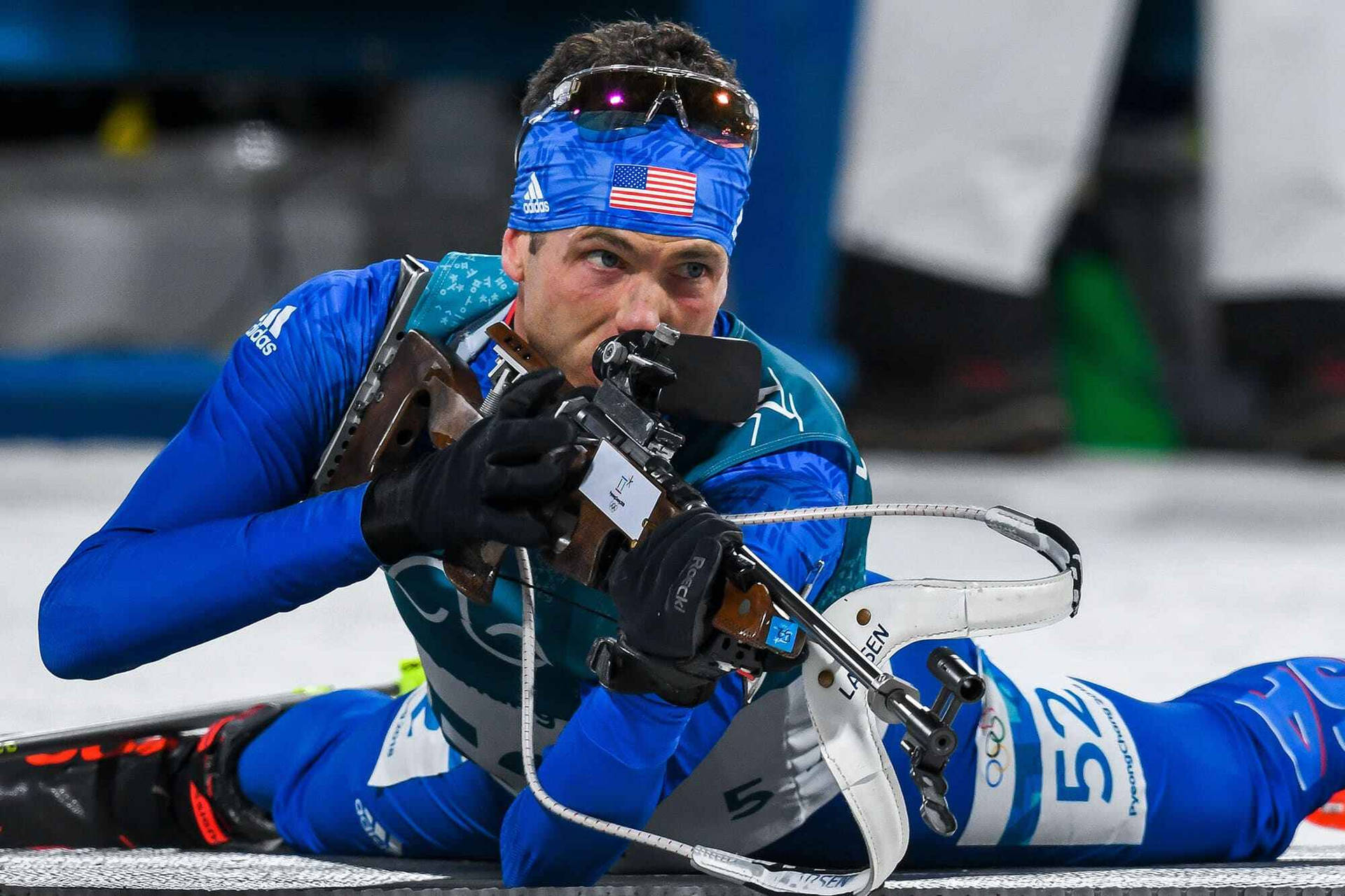 2018年平昌冬季奧運會美國越野滑雪射擊運動員提姆·伯克 壁紙 — 一位專注的美國冬季兩項運動員身穿藍色裝備，在雪地地形上的冬季比賽中瞄準步槍。