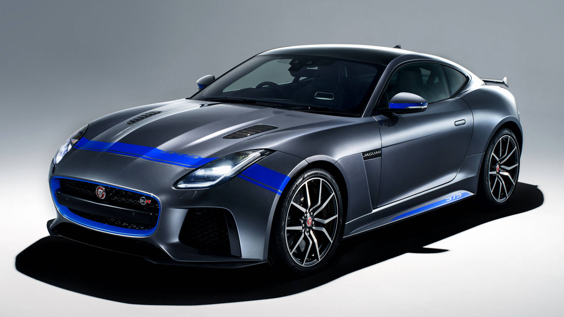 2018款捷豹F-Type圖 壁紙 — 一輛時尚的銀色 Jaguar F-Type 跑車，帶有醒目的藍色賽車條紋和黑色輪圈，以極簡灰色工作室背景為襯托。