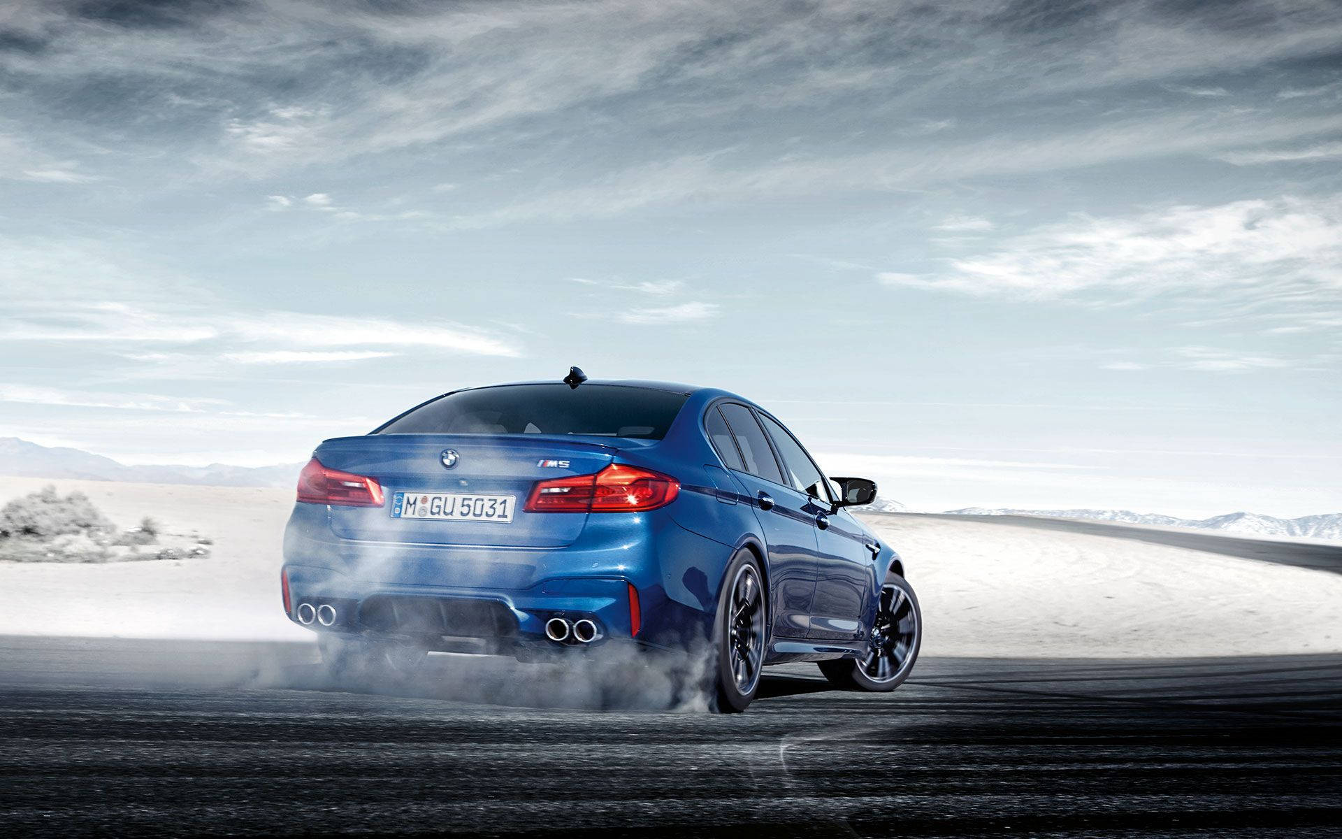 2019 BMW M5系列 壁紙 — 一輛鮮豔的藍色BMW M5在陰天下的黑暗賽道上漂移，輪胎冒出滾滾濃煙，背景是廣闊的雪沙漠。