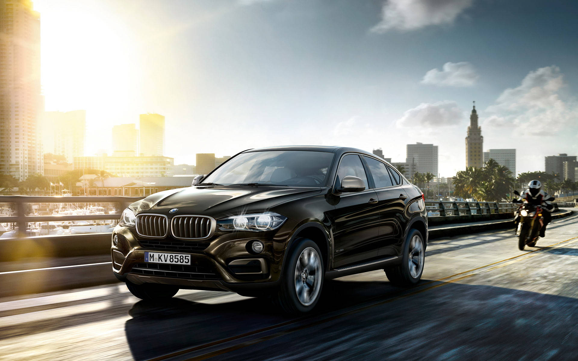 2019款BMW X6 M 壁紙 — 一架時尚的黑色BMW X6 SUV在陽光明媚的橋上行駛，後面有一位電單車騎士，背景是城市天際線。
