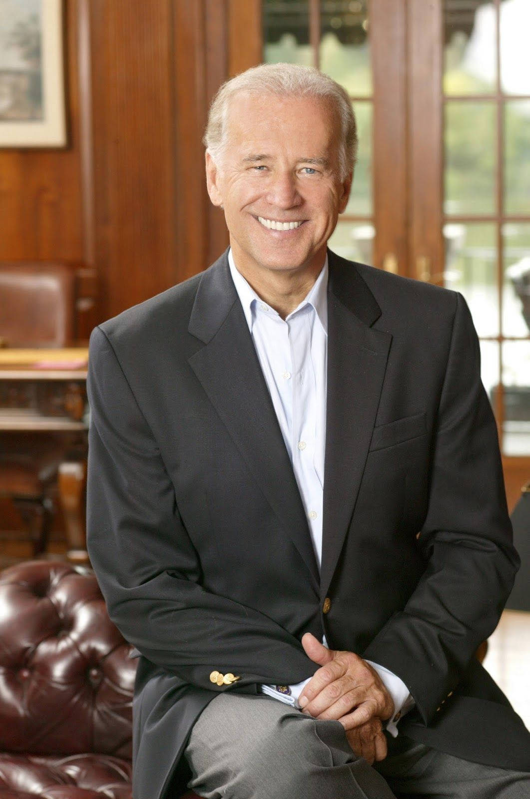 喬·拜登2020競選期間微笑 壁紙 — 一個微笑的男人穿著深色西裝，自信地坐在一個有木鑲板房間的紅色皮革簇絨椅上，旁邊有大窗戶。