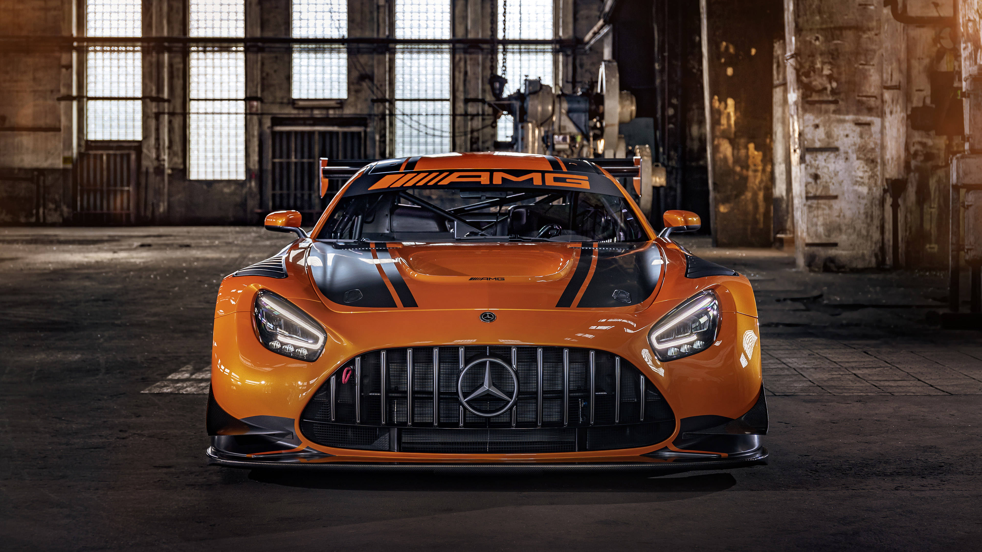 2020 年梅賽德斯-AMG GT R Pro 頂級展示 壁紙 — 一輛充滿活力的橙色Mercedes-AMG GT3賽車在一個有高窗和混凝土地板的骯髒工業倉庫中面向前方。