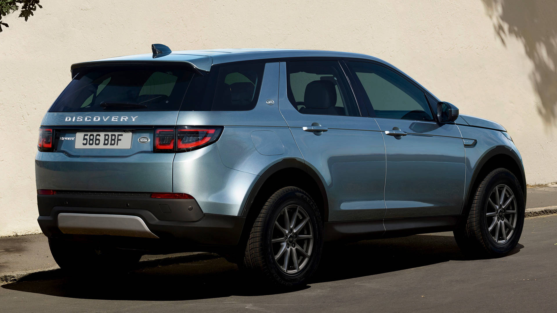 2020 路虎發現運動版 iPhone 主題壁紙 壁紙 — 一架時尚的淺藍色 Land Rover Discovery SUV 以傾斜角度停泊在陽光照射的米色牆壁旁，投射出銳利的陰影。