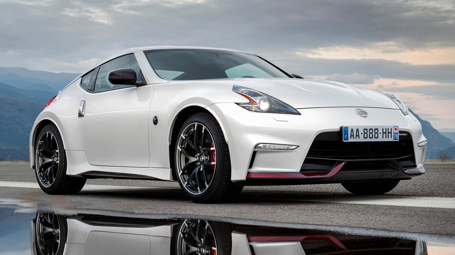 2020尼桑GT-R Nismo動態展示 壁紙 — 一架線條流暢的白色日產370Z跑車，配備黑色輪圈和紅色點綴，停在潮濕的道路上，在陰雲密佈的山景背景下反射出光澤的表面。