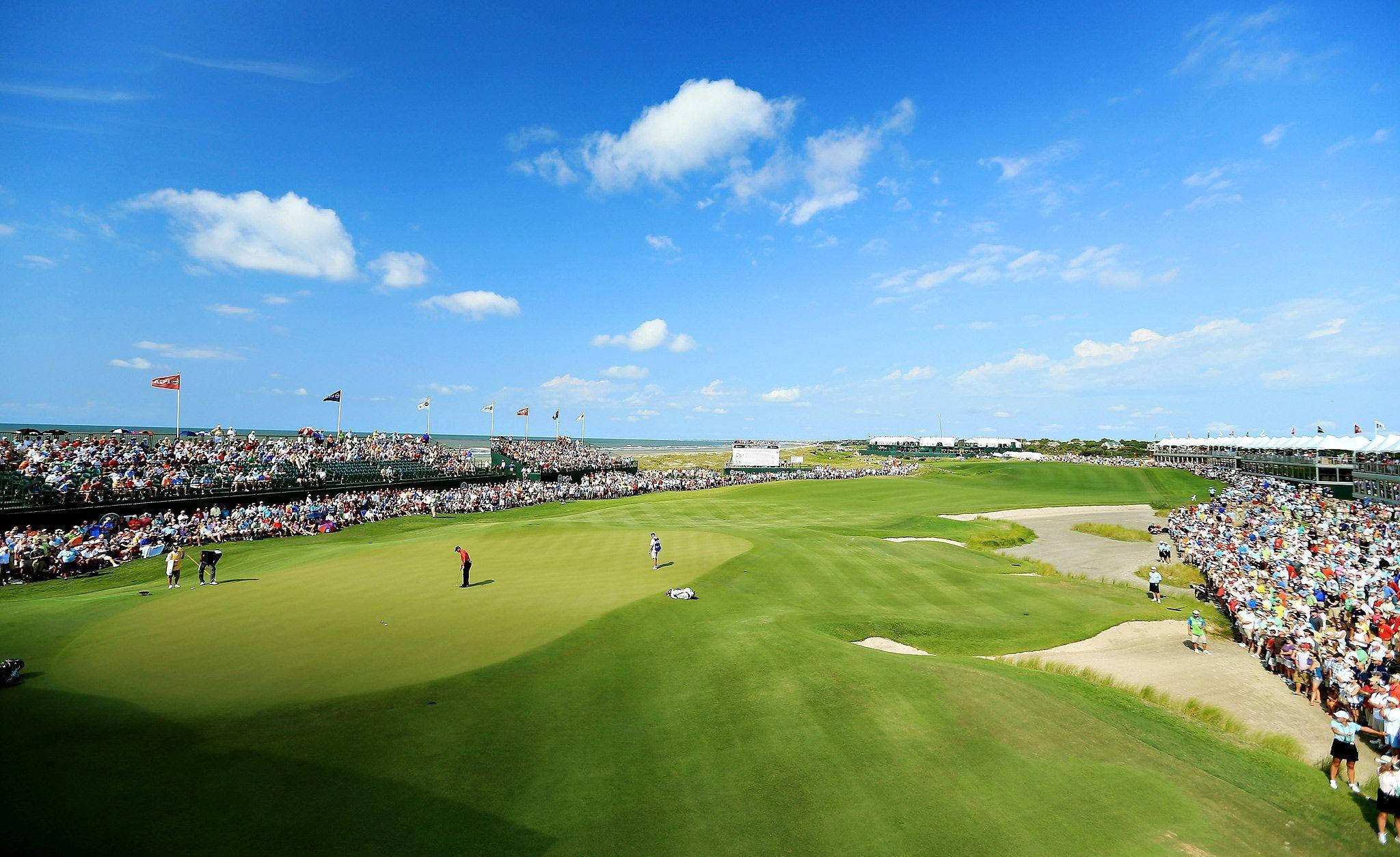 2021 PGA 比賽 基亞瓦島 壁紙 — 在高空俯瞰一個充滿活力的綠色高爾夫球場，上方是明亮的藍天，散落著白雲，觀眾看台座無虛席。