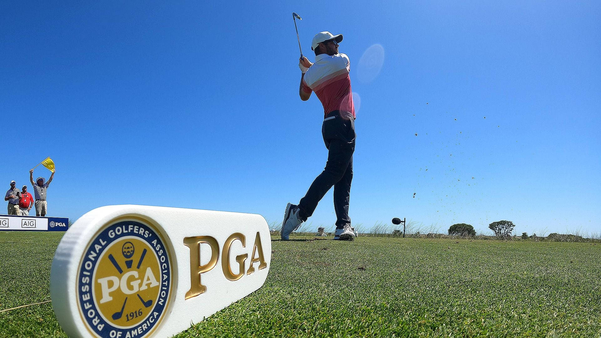 2021 PGA 錦標賽 記號牌 壁紙 — 一名高爾夫球手在陽光明媚的球道上強力揮桿，前景有PGA標誌標記，背景有觀眾歡呼。