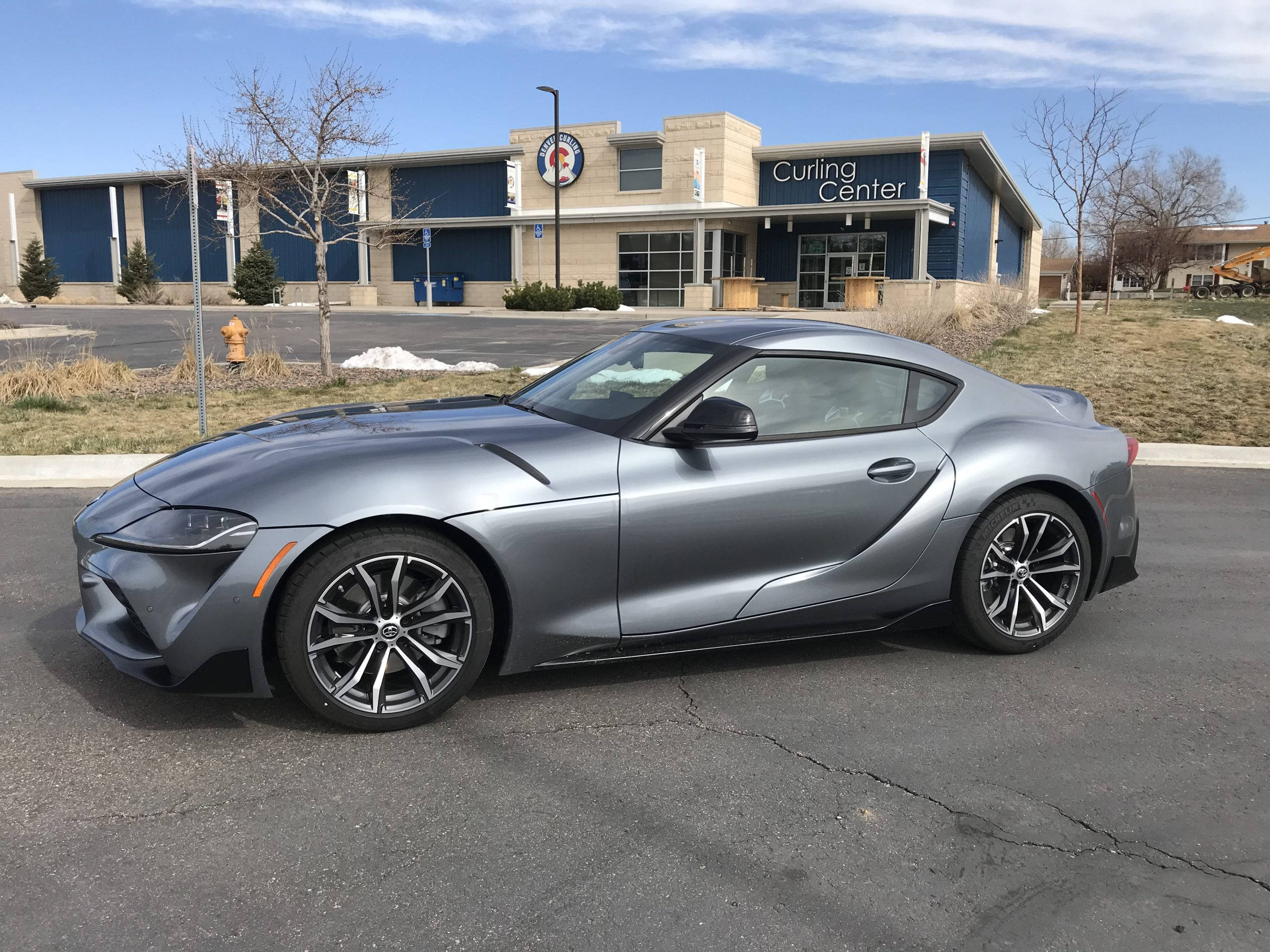 2021 Toyota Supra 2.0 壁紙 — 一架時尚的銀色跑車停泊在冰壺中心前，天氣晴朗，樹木光禿，地上有積雪。