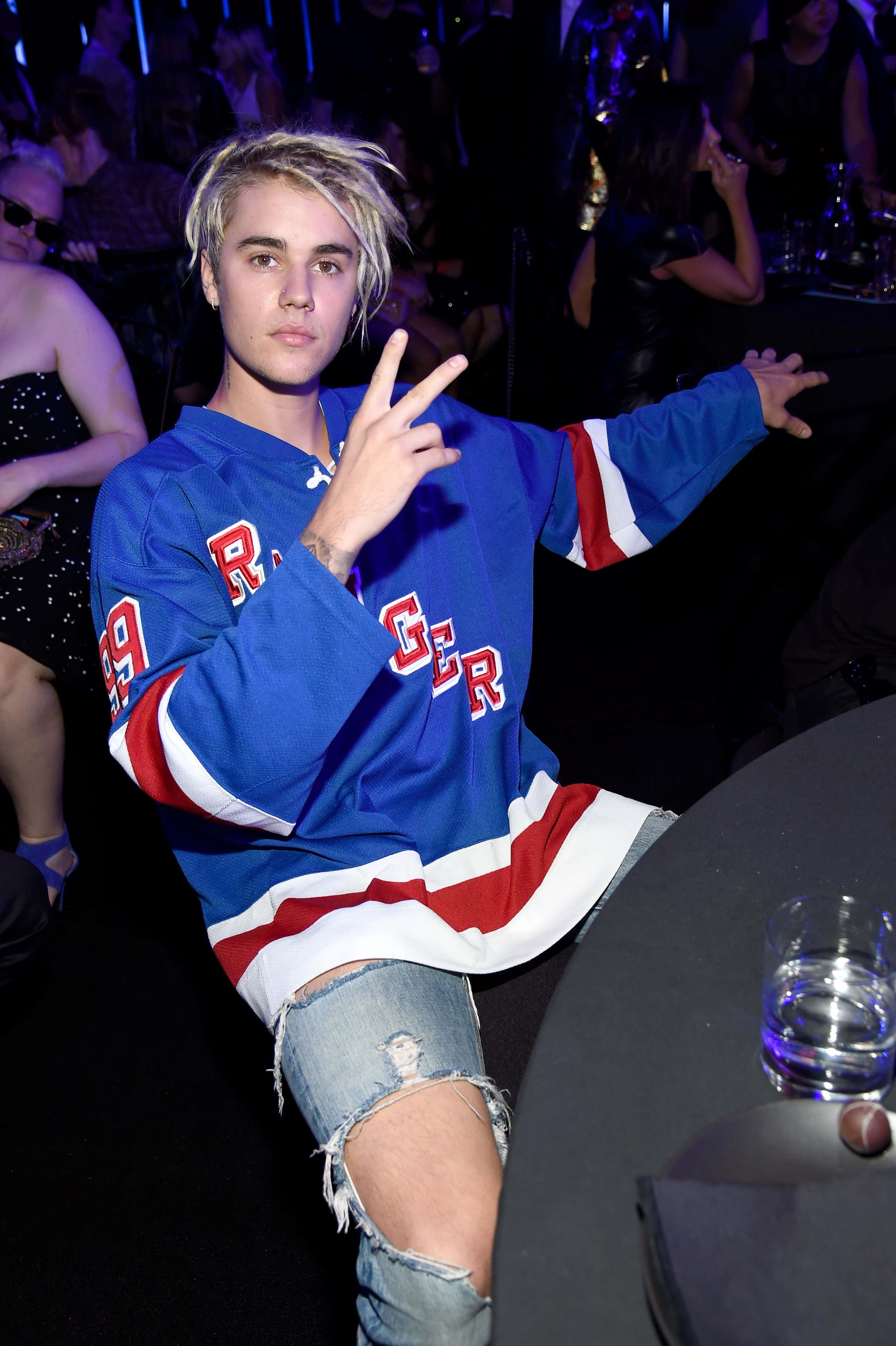 2021年賈斯汀·比伯的時尚造型 壁紙 — Justin Bieber 自信地擺姿勢，身穿藍、紅、白色冰球球衣，在燈光昏暗的派對中，背後有閃爍的燈光和人群。