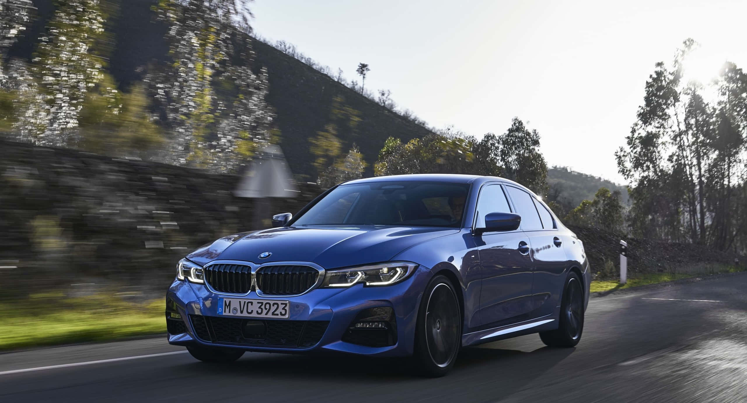 2021款BMW 330i車型壁紙 壁紙 — 一輛時尚的藍色寶馬轎車在明亮的日光下，沿著蜿蜒的山路疾馳，綠樹和石牆模糊成一片。