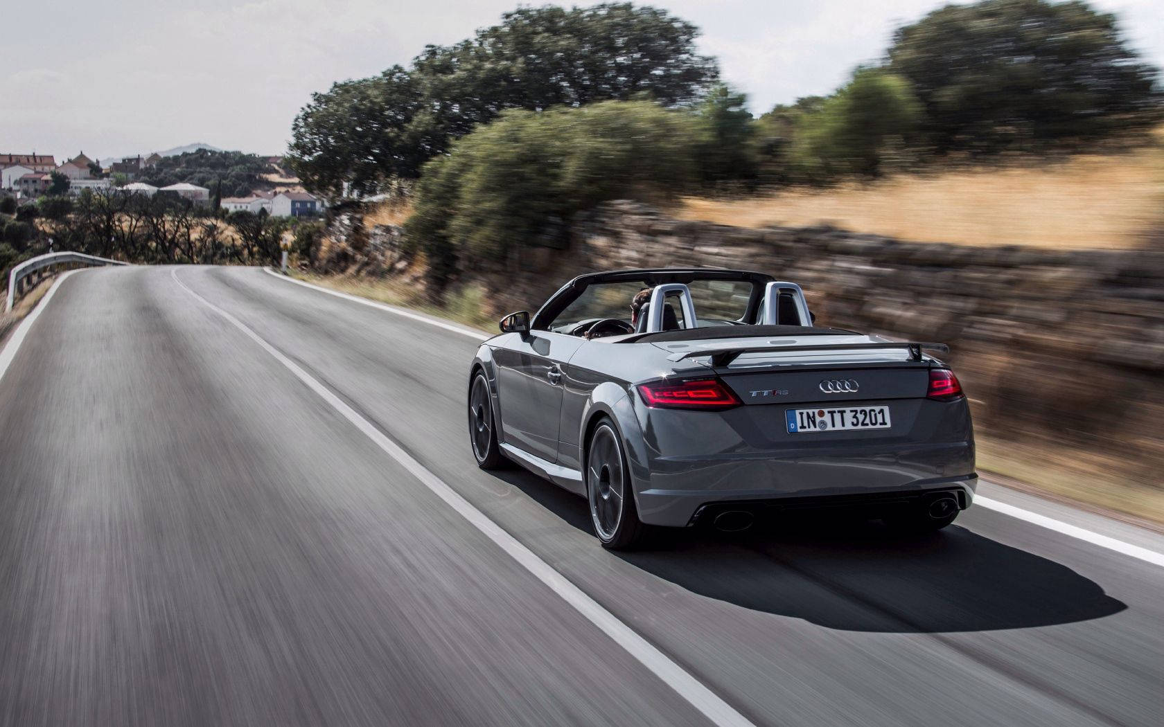 灰色Audi TT Roadster在彎曲的鄉村道路上快速行駛，背景有樹木和房屋。