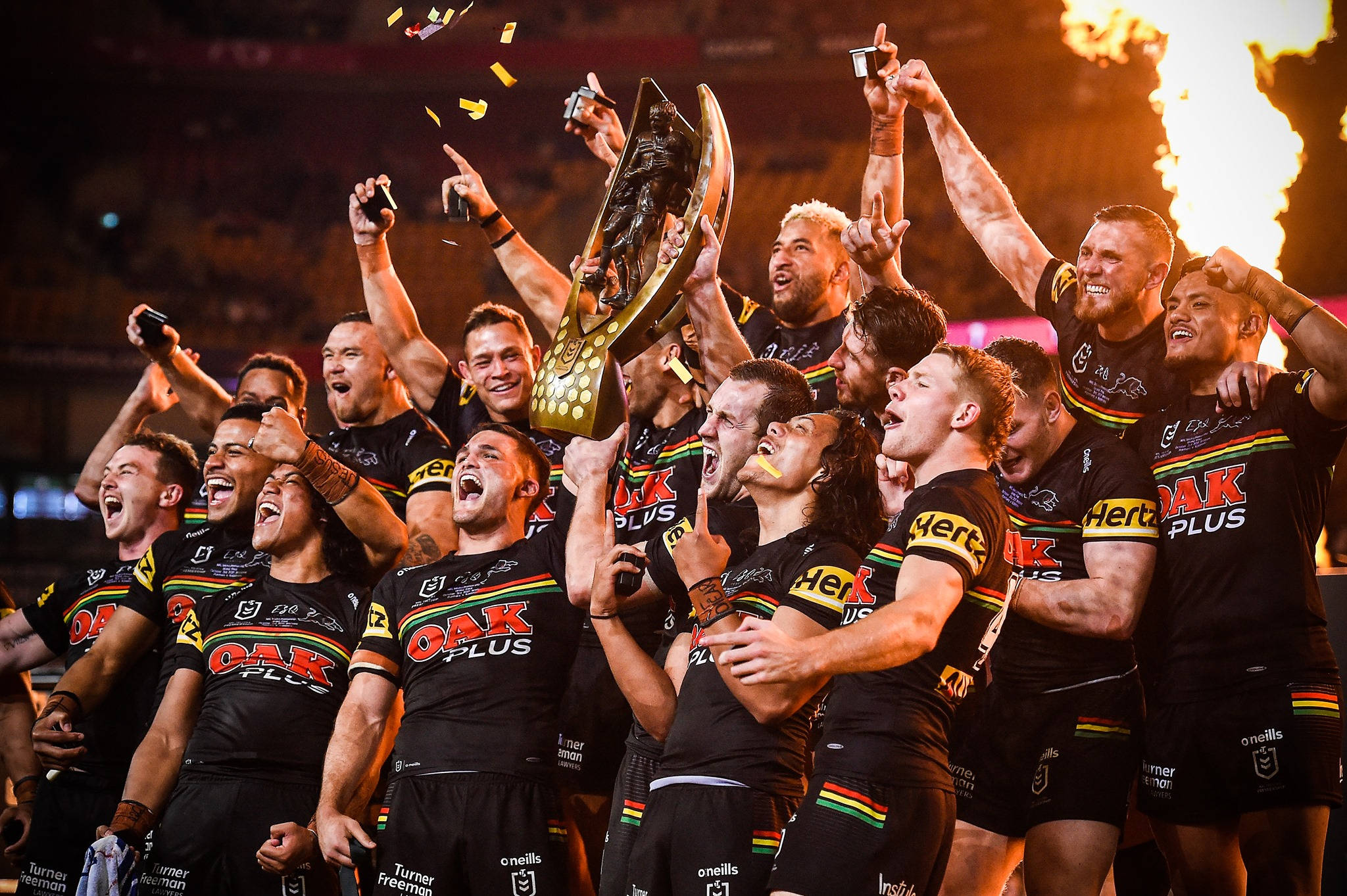 2021年佩斯頓黑豹隊NRL 壁紙 — 身穿黑色球衣的橄欖球聯賽球員瘋狂慶祝，手持金色獎盃，彩紙紛飛，身後火焰噴射。